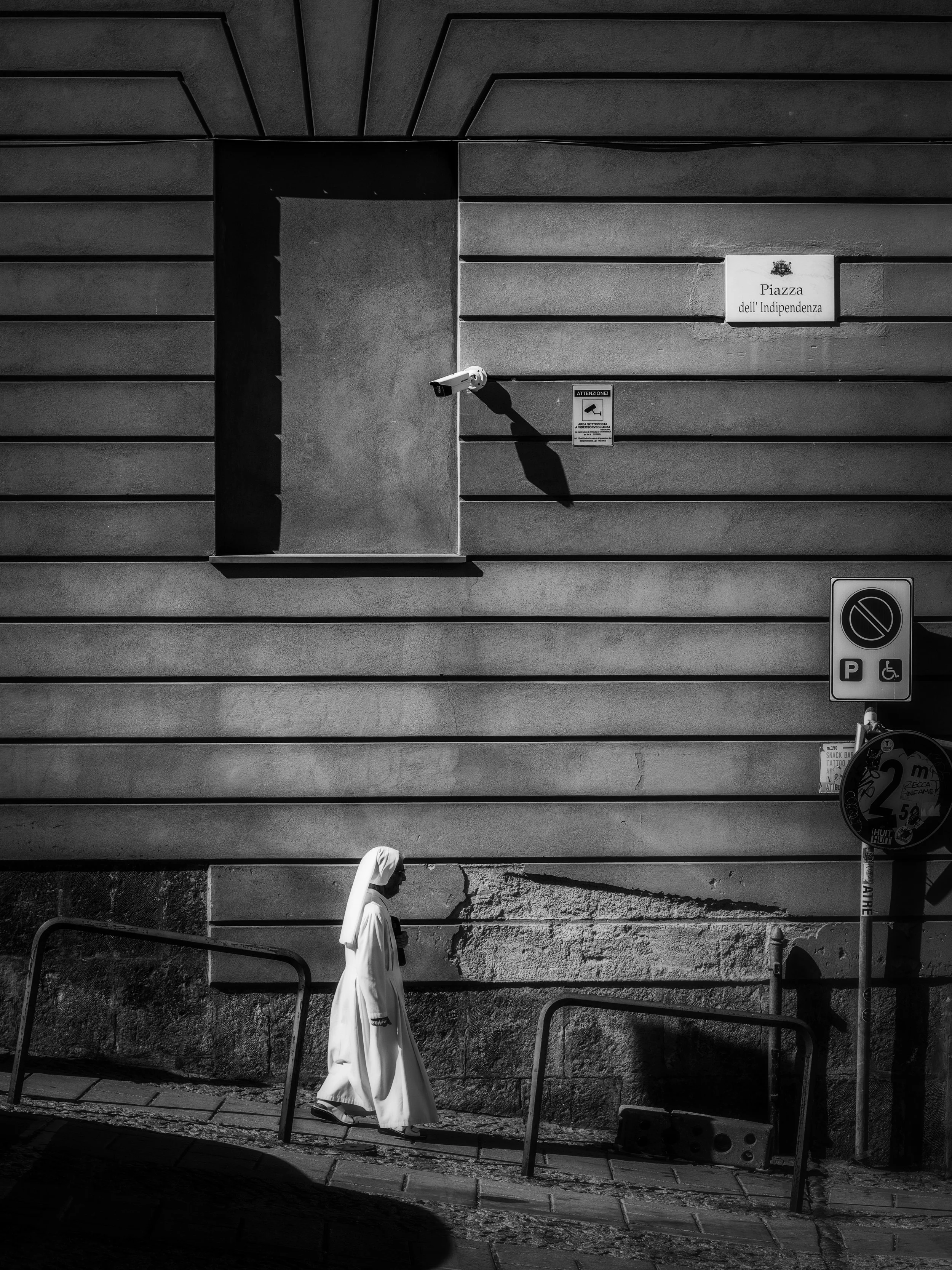 A person dressed in white clothing walking along a sidewalk next to a wall with horizontal lines. There are signs, a security camera, and a parking sign on the wall. The scene is in black and white.