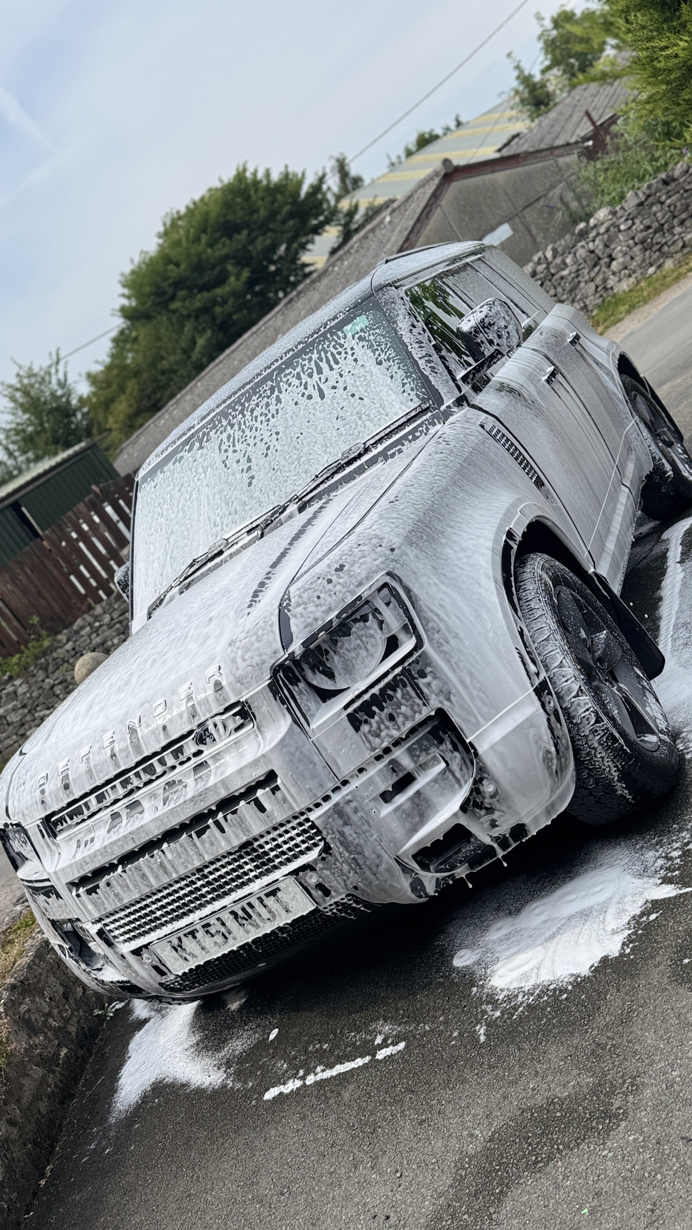 Silver Range Rover covered in soap suds during washing, on a driveway with soap and water on the ground.