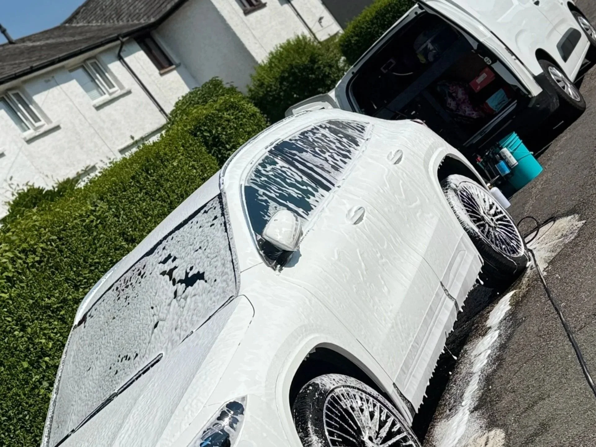 White car covered in foam soap with a black car behind it and a green hedge in the background.