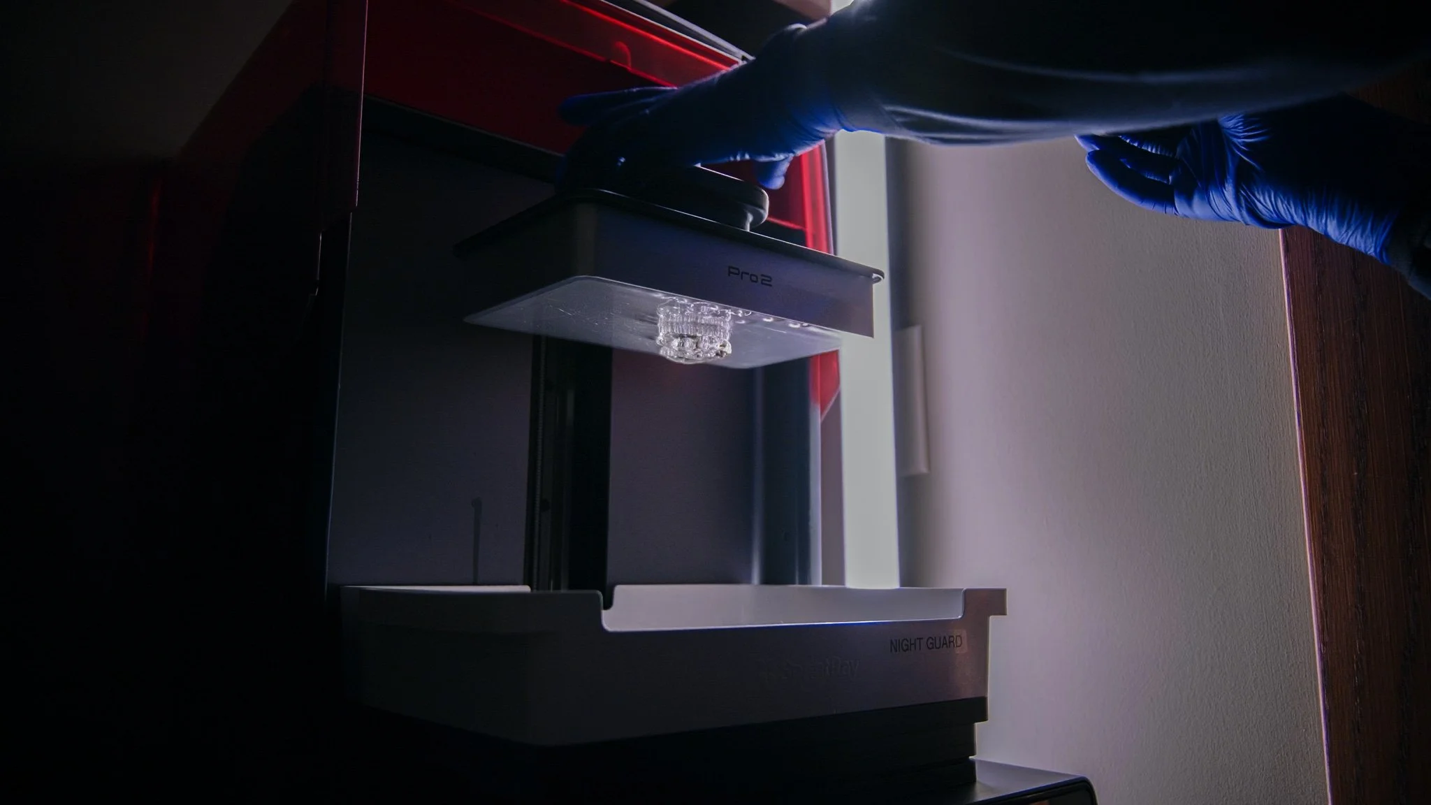 A laboratory setting with a person wearing blue gloves handling a small transparent bottle under a machine labeled 'Pro 2.' The scene is dimly lit with a focus on the person's hands and the equipment.