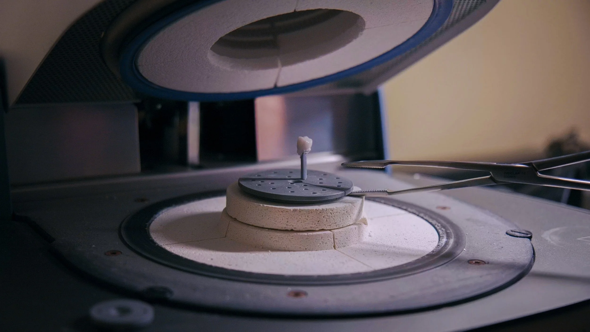 Close-up of a dental milling machine with a small dental prosthetic held by tweezers inside the machine.