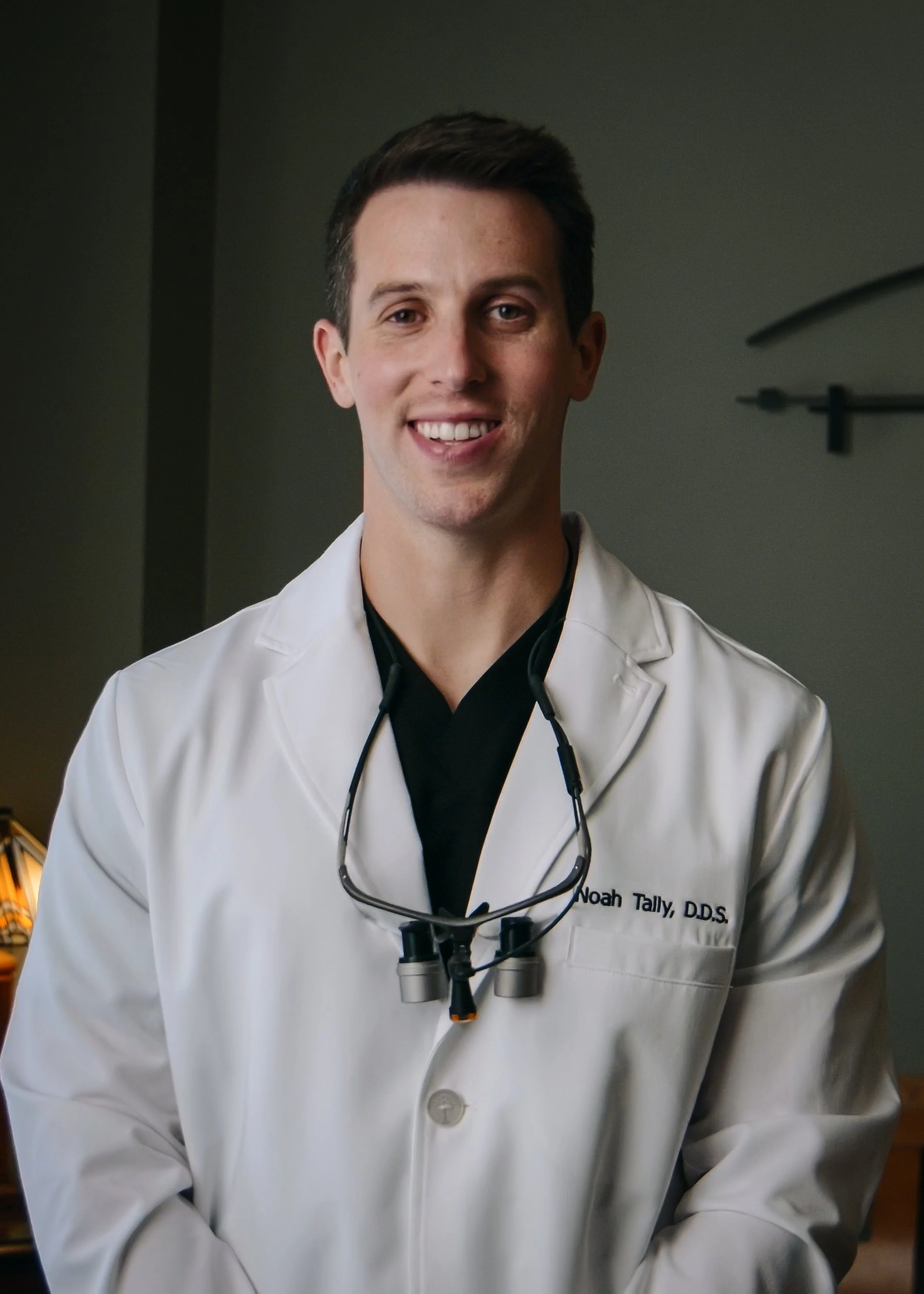 A smiling man with short dark hair and light skin, wearing a white medical coat with a name tag that reads Noah Tally, DDS, and a black shirt underneath. He has dental loupes hanging around his neck, standing in a professional setting.