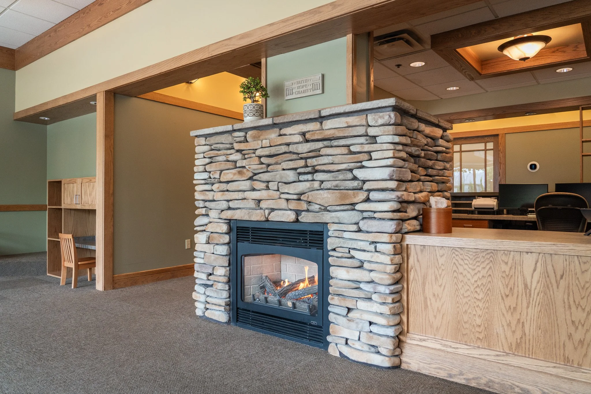 Interior of a building with a stone fireplace, wooden accents, a ceiling light, and office furniture visible in the background.