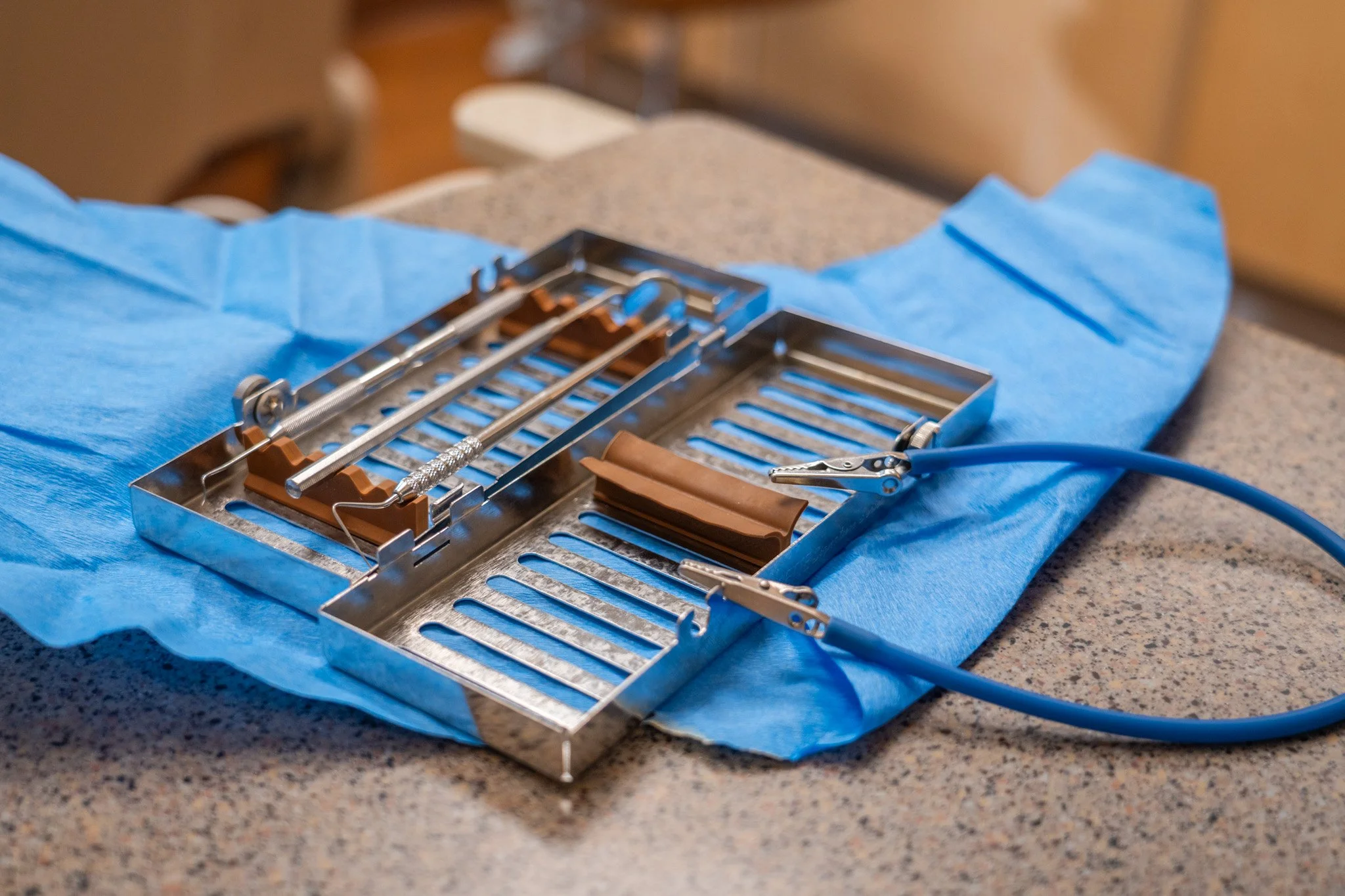 A dental surgical retractor, placed on a blue surgical drape on a speckled counter, with dental tools and brown rubber dental impression material on it
