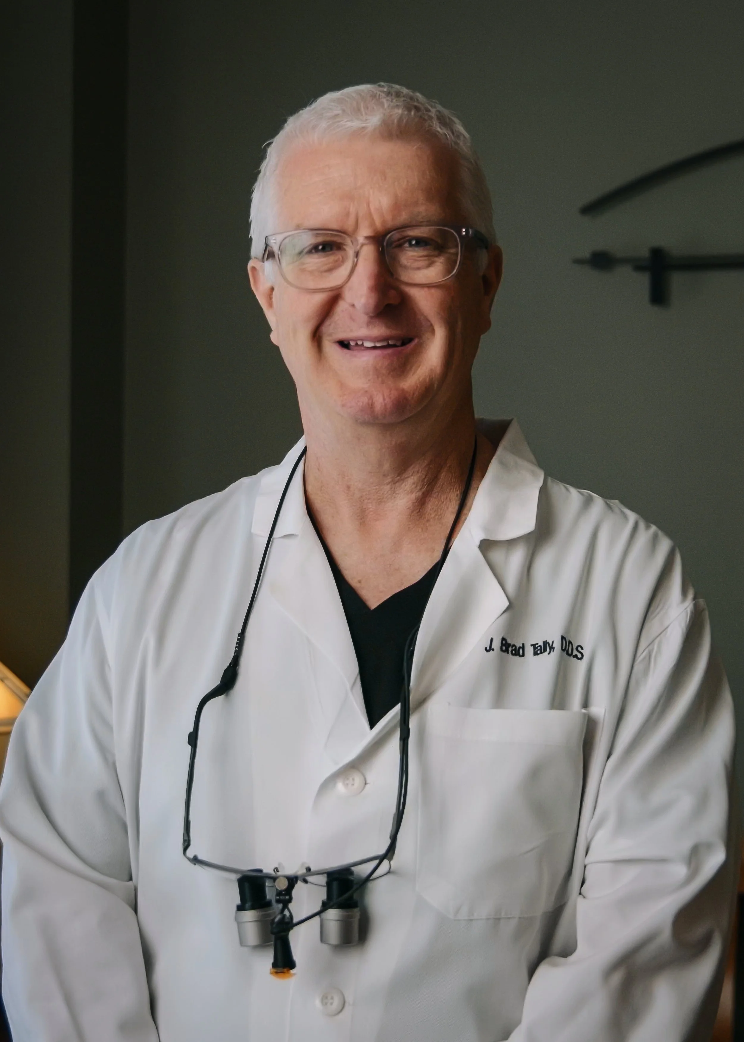 A smiling man wearing glasses and a white lab coat with a name tag that reads 'J. Brad Tally DDS'. He has a stethoscope around his neck and orthodontic loupes hanging from his neck. The background is a dark greenish wall with a clock partially visible on the right.