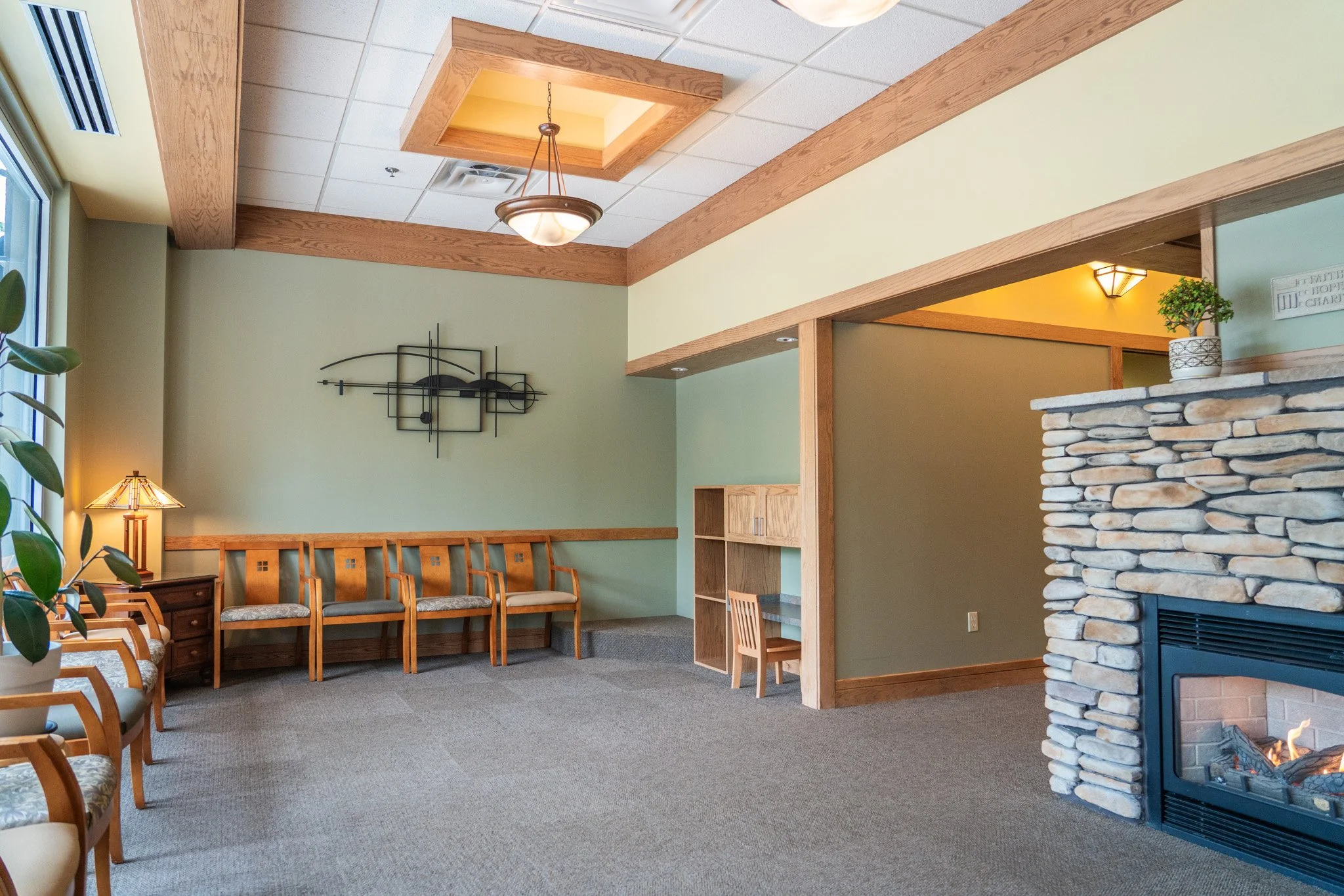 Interior of a waiting room with wooden chairs, a stone fireplace, a decorative wall art piece, and a window with natural light, featuring a ceiling with wooden beams and modern lighting fixtures.