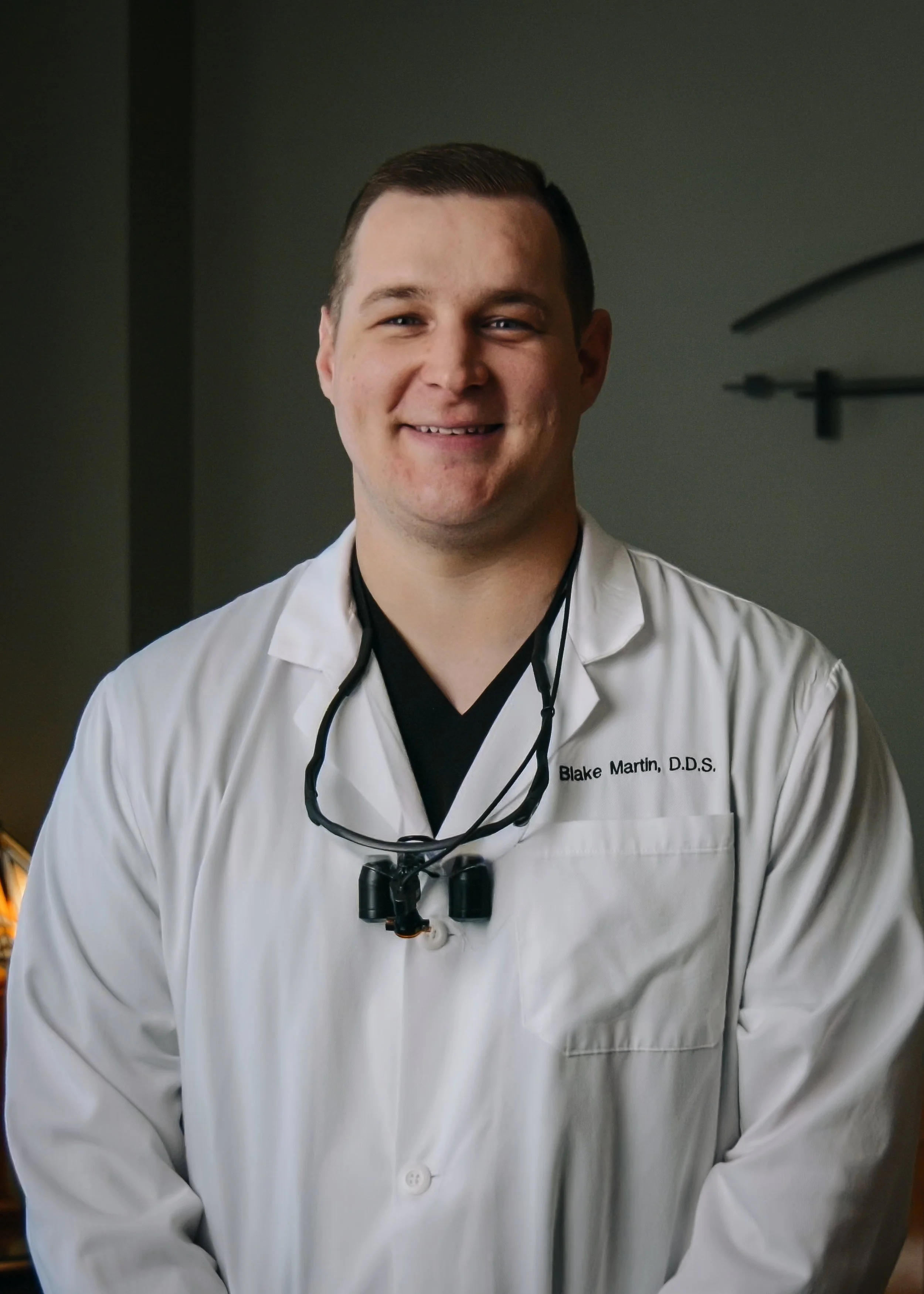 A young man with light skin and short dark hair, wearing a white medical coat with the name tag 'Blake Martin, D.D.S.' and a black shirt underneath. He has a stethoscope around his neck and small glasses hanging from it, smiling in a medical or dental office setting.