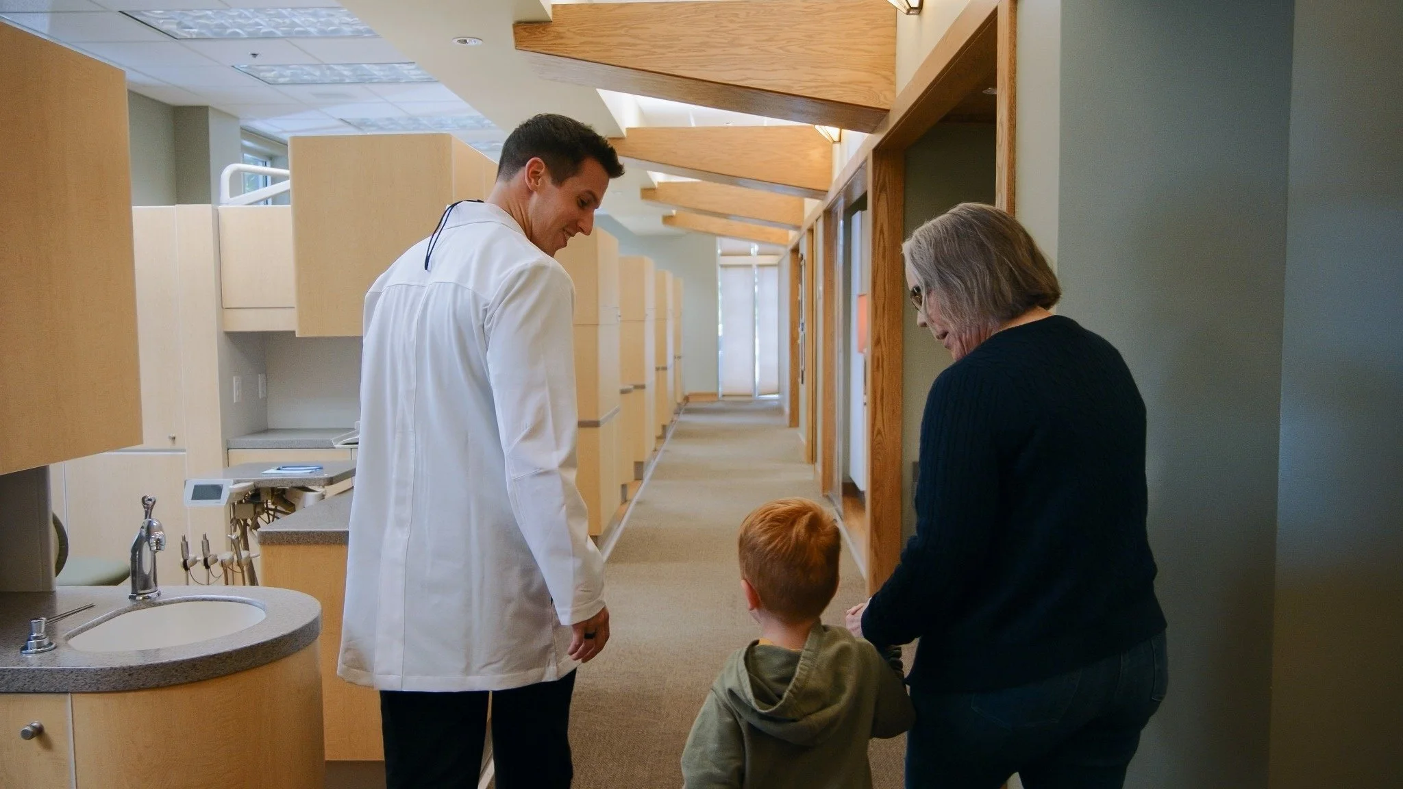 A healthcare professional, a young man in a white lab coat, interacts with a woman and two children in a medical facility hallway, with wooden accents and multiple exam rooms.