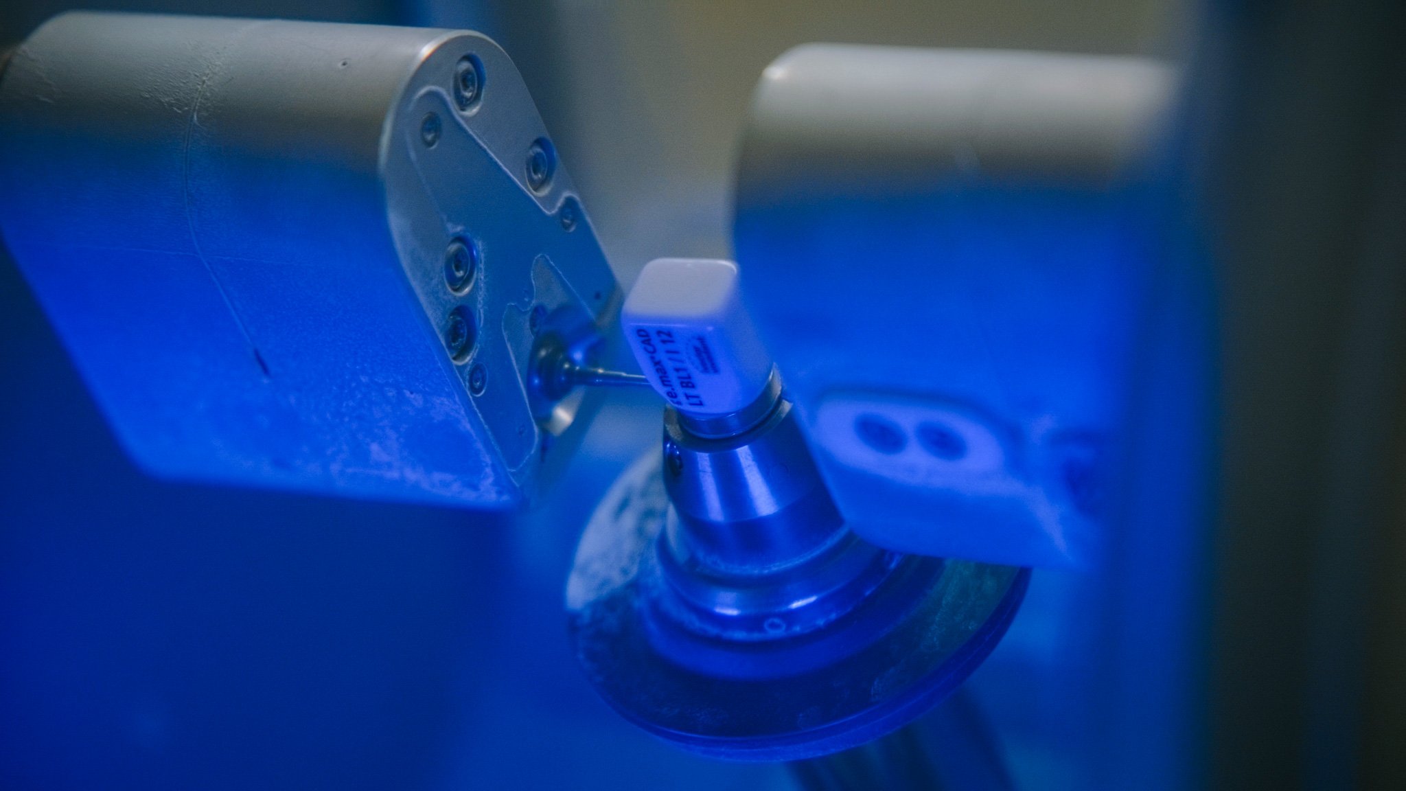 Close-up of a scientific instrument or piece of laboratory equipment, illuminated with blue light, showing metal parts and a white component attached to a metal base.