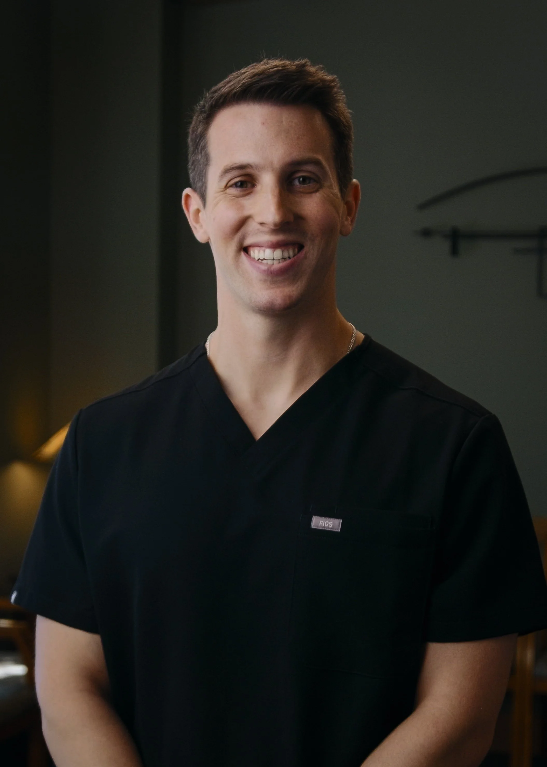 Close-up of a smiling man wearing a black shirt with a small patch that says 'FIGS', standing indoors with a dark background and blurred furniture.