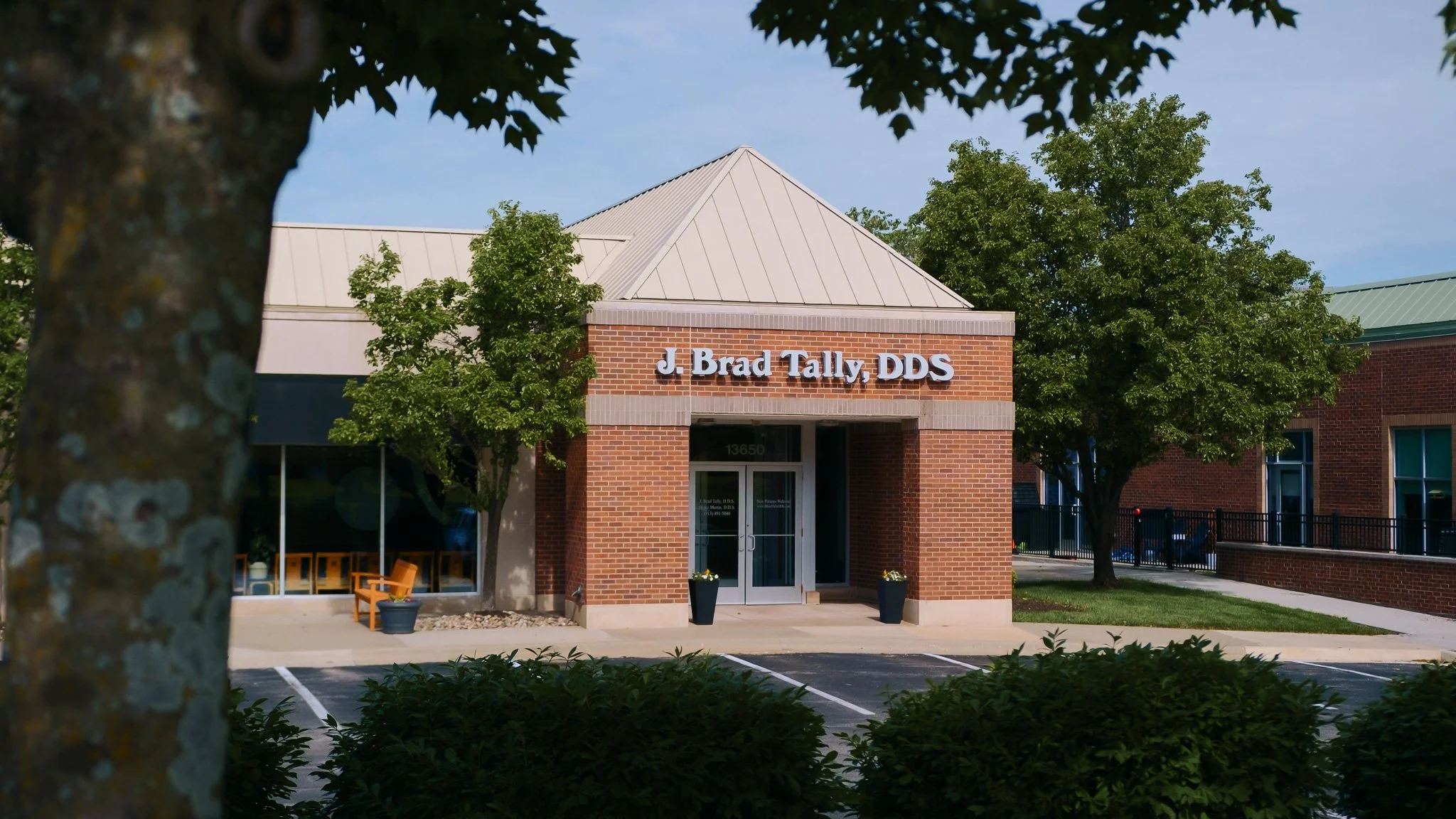 Exterior view of a dental office building with a brick facade and signage that reads 'J. Brad Tally, DDS.' There are trees and parking spaces in front of the building, with some outdoor seating visible through the windows.