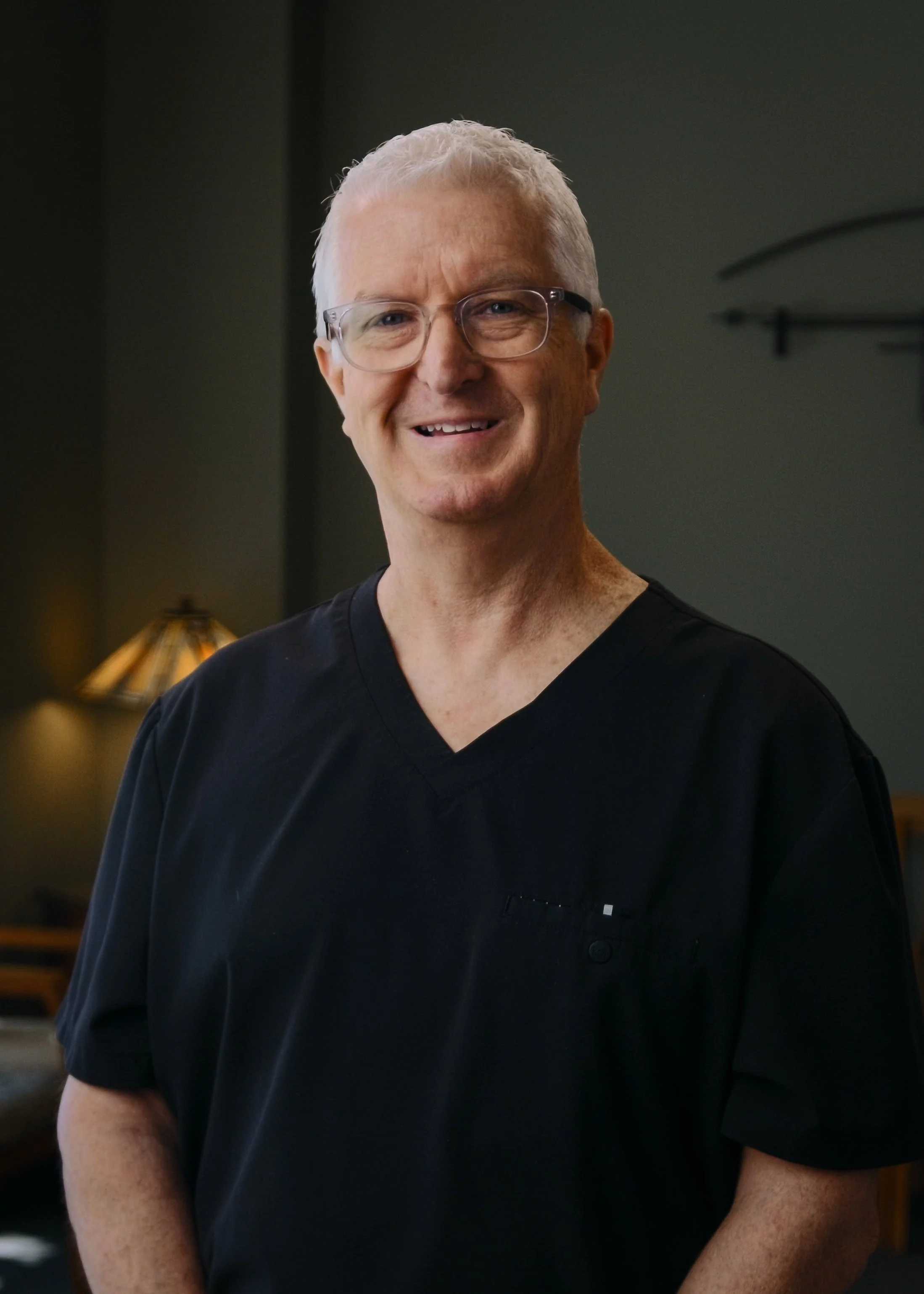 A middle-aged man with short gray hair and glasses smiling, wearing a black medical scrub top, indoors with dim lighting and a lamp in the background.