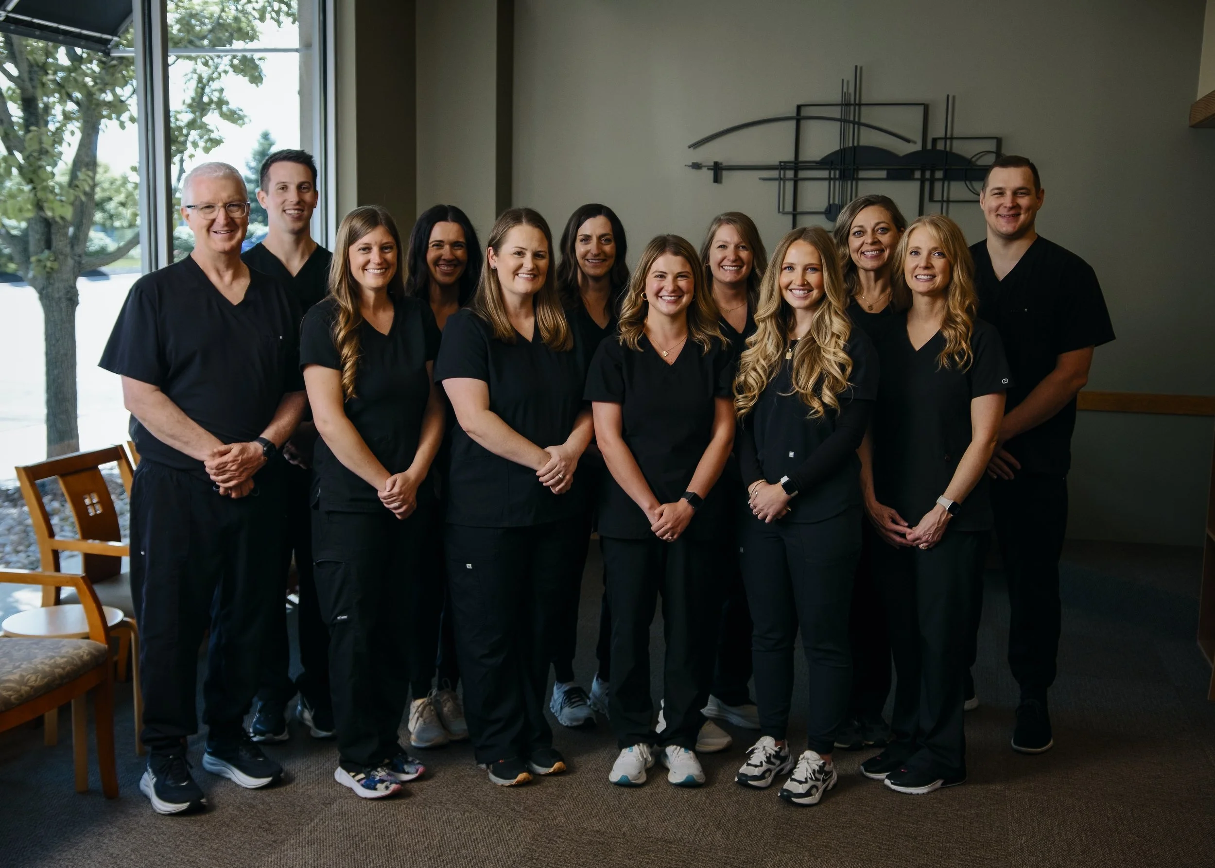 Group of eleven healthcare professionals wearing black scrubs posing indoors, with large windows and a metal wall art piece in the background.
