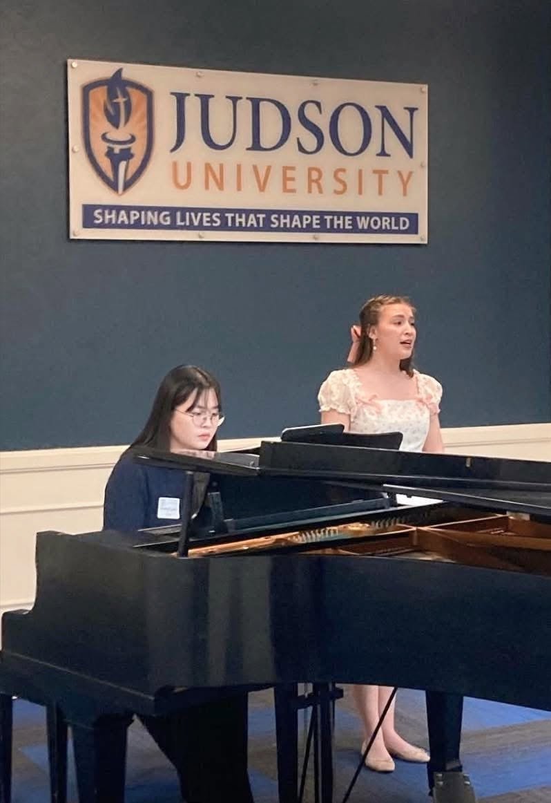 A woman singing and a woman playing piano in a room with a Judson University sign on the wall.