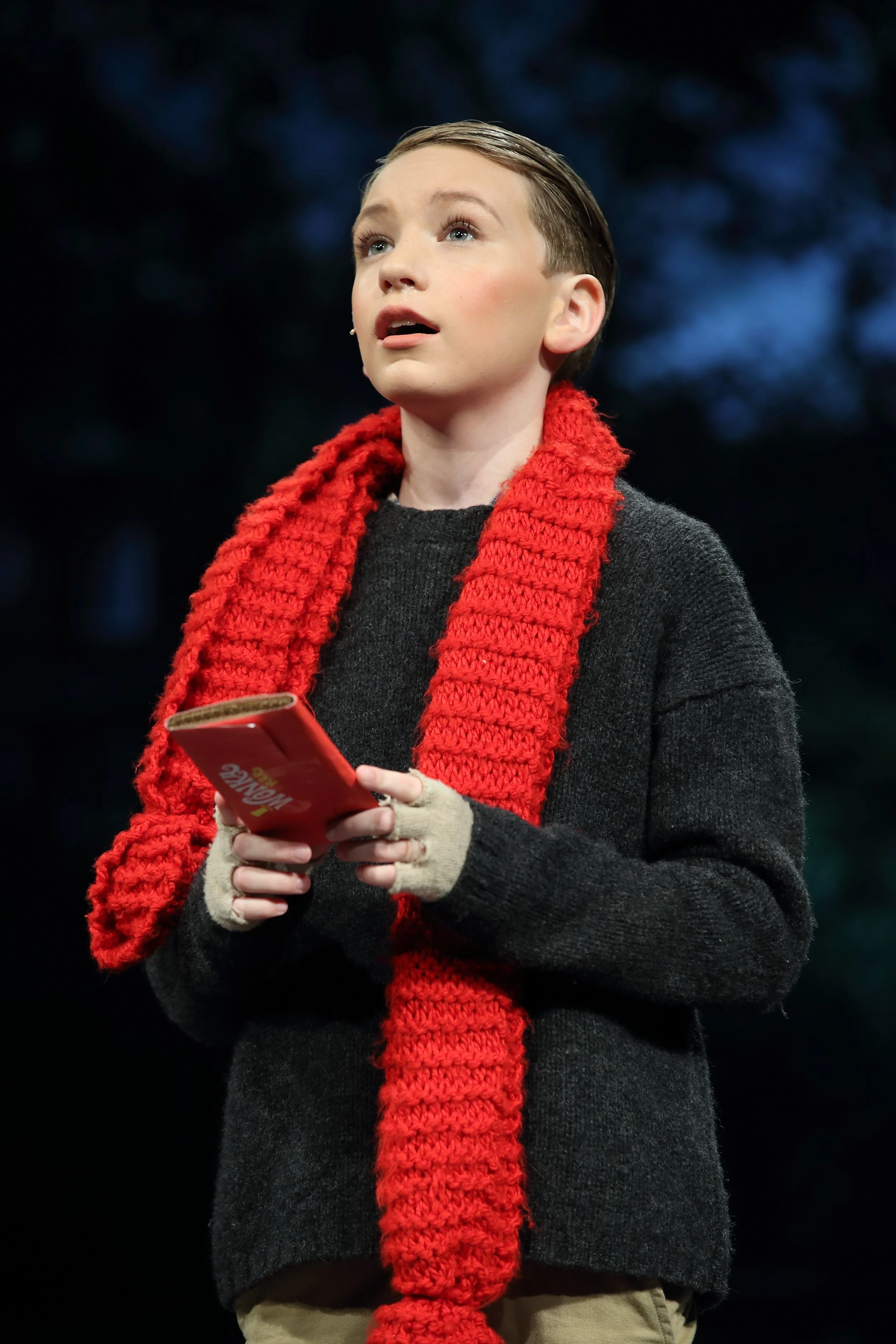 A young person with short hair, wearing a red scarf, black sweater, and beige pants, holds a red book and looks upward with an expression of wonder against a dark, out-of-focus background.