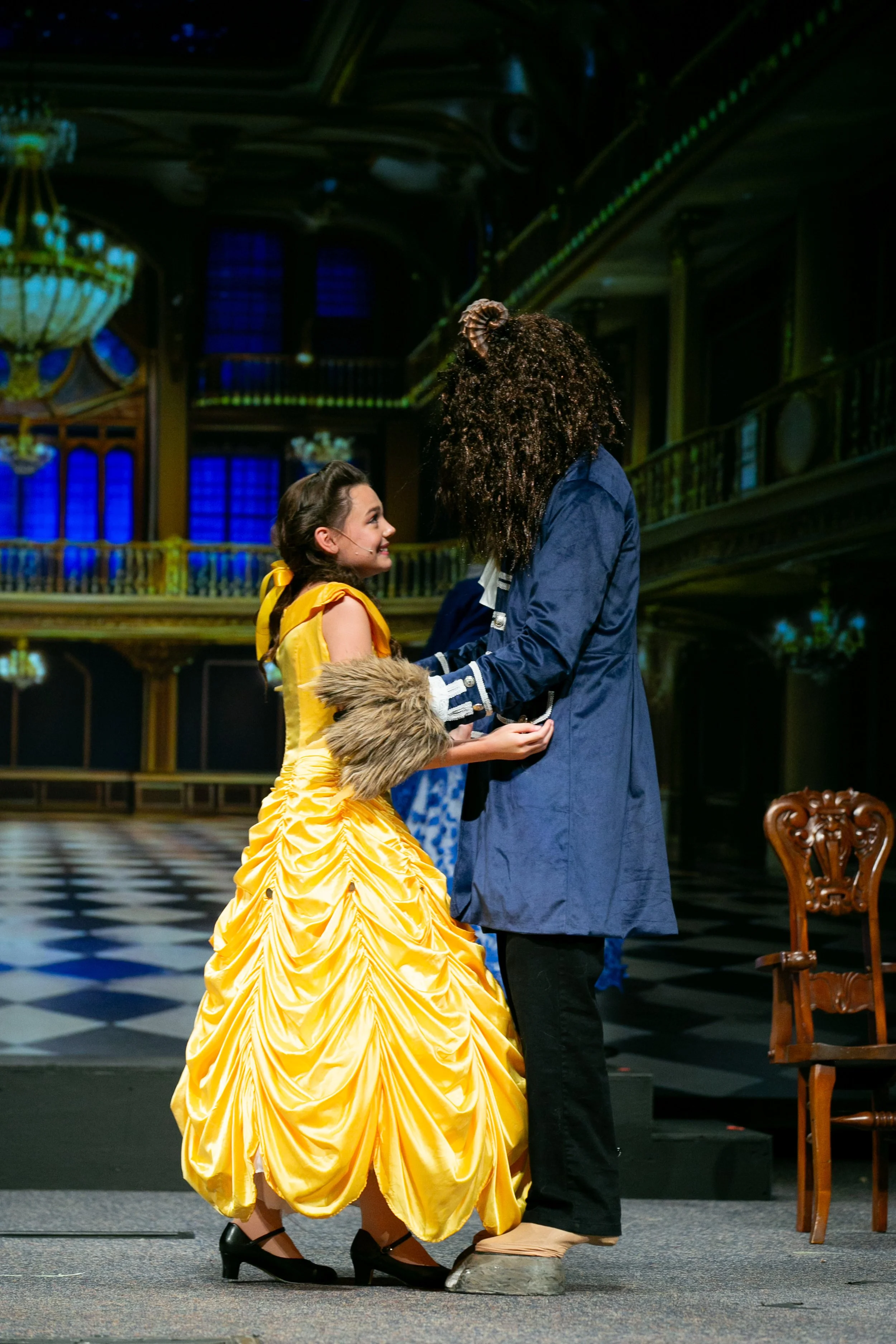 A woman in a yellow ball gown and a man in a blue coat singing together on stage, holding hands, in a theater setting.