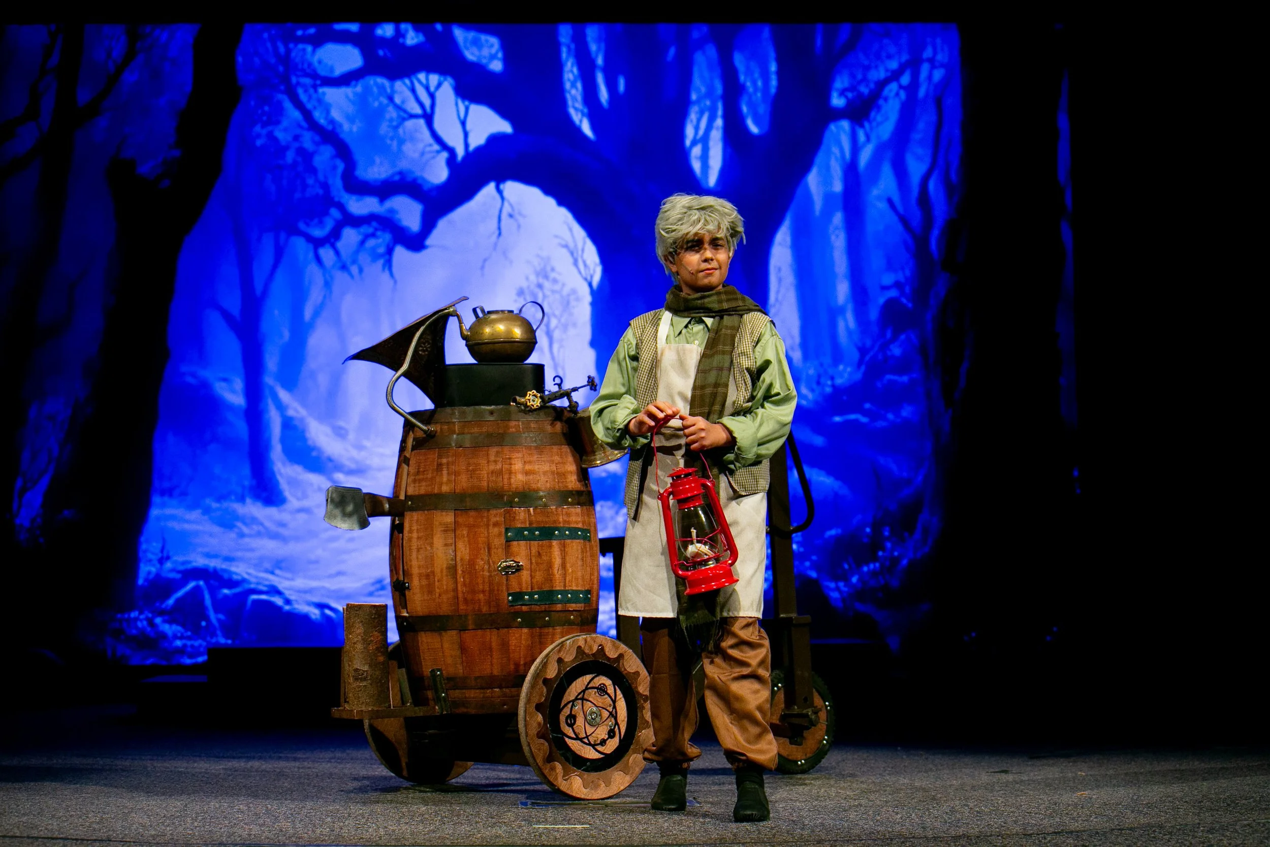 A young boy dressed as an inventor or scientist, standing on a stage with a blue forest background. He holds a red lantern in one hand and is near a wooden contraption with various gears and a teapot on top.
