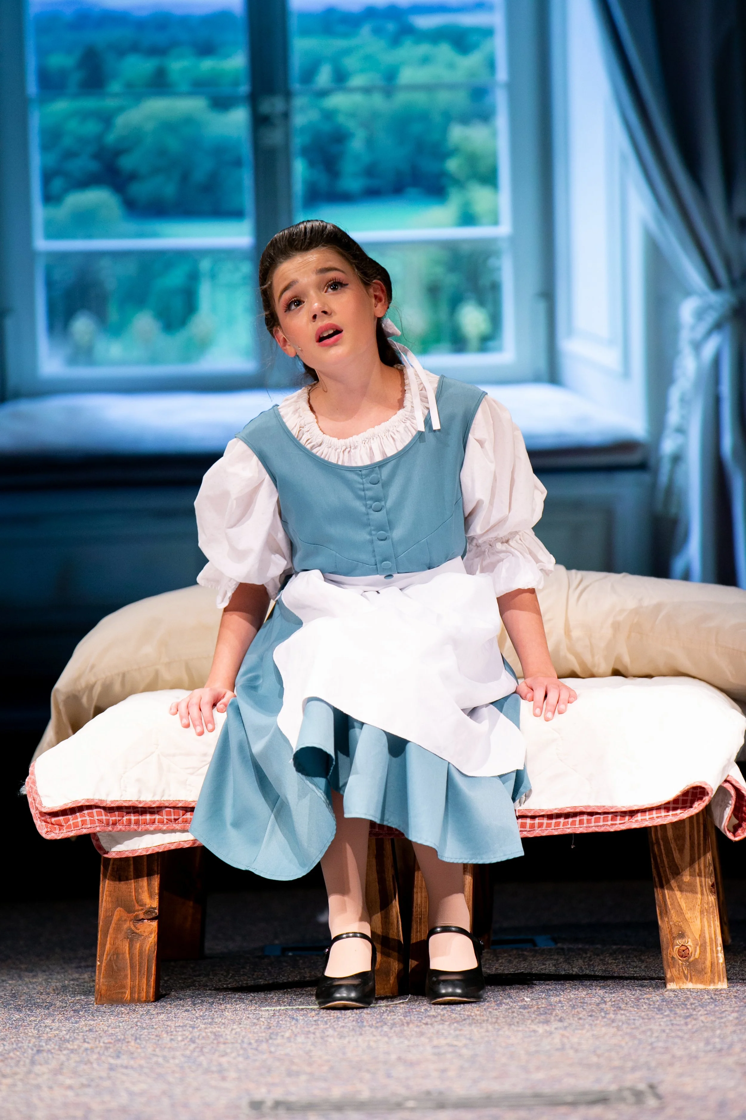 Young girl dressed in historical clothing, sitting on a wooden bench with her legs hanging over the edge, in a room with large window showing green landscape outside, suggesting a period-piece or theatrical setting.