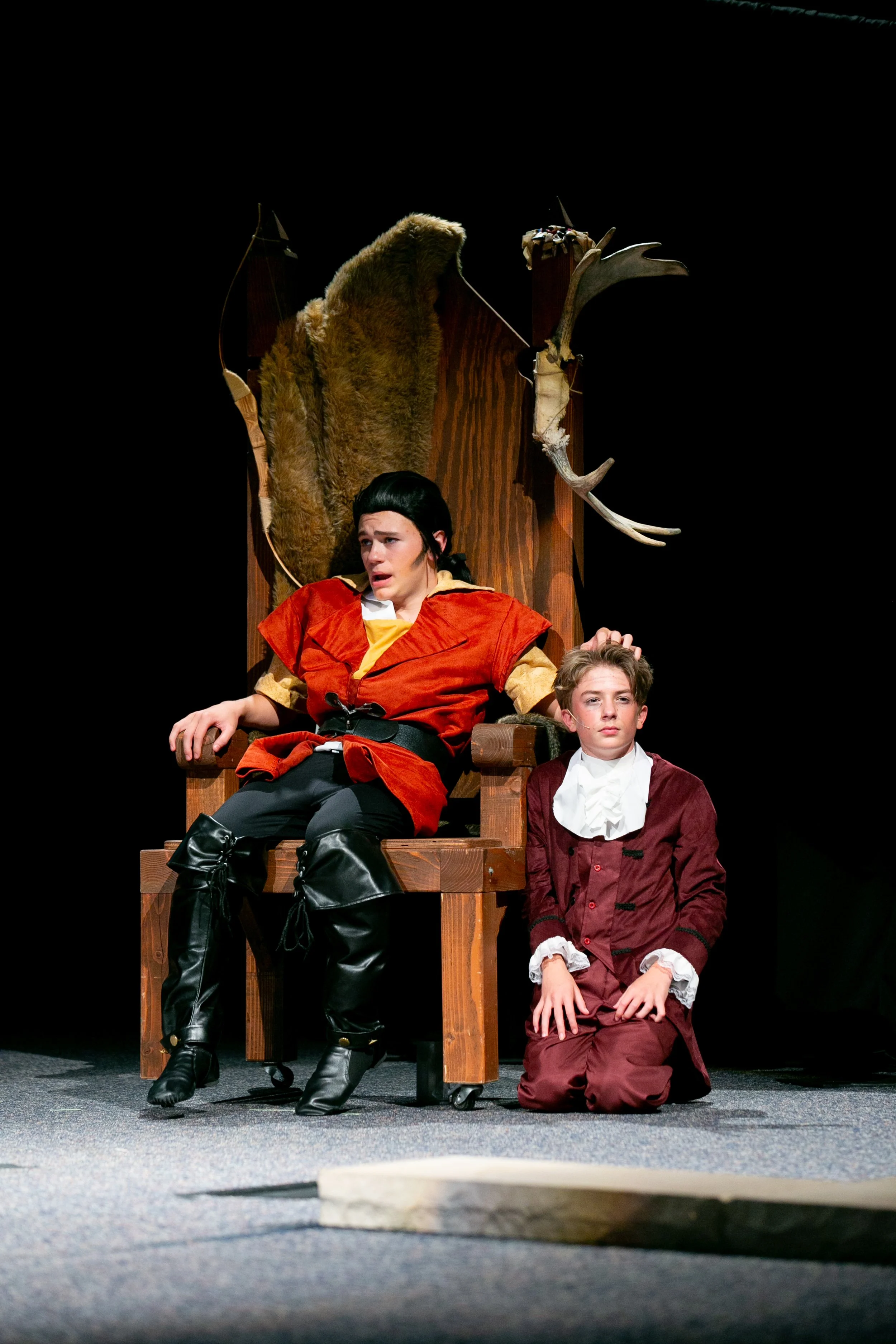 Two young boys dressed in historical costumes sit on a stage with a wooden throne and mounted animal skulls behind them. One boy has dark hair and is dressed in a red and yellow costume, sitting on the throne looking distressed. The other boy has lig