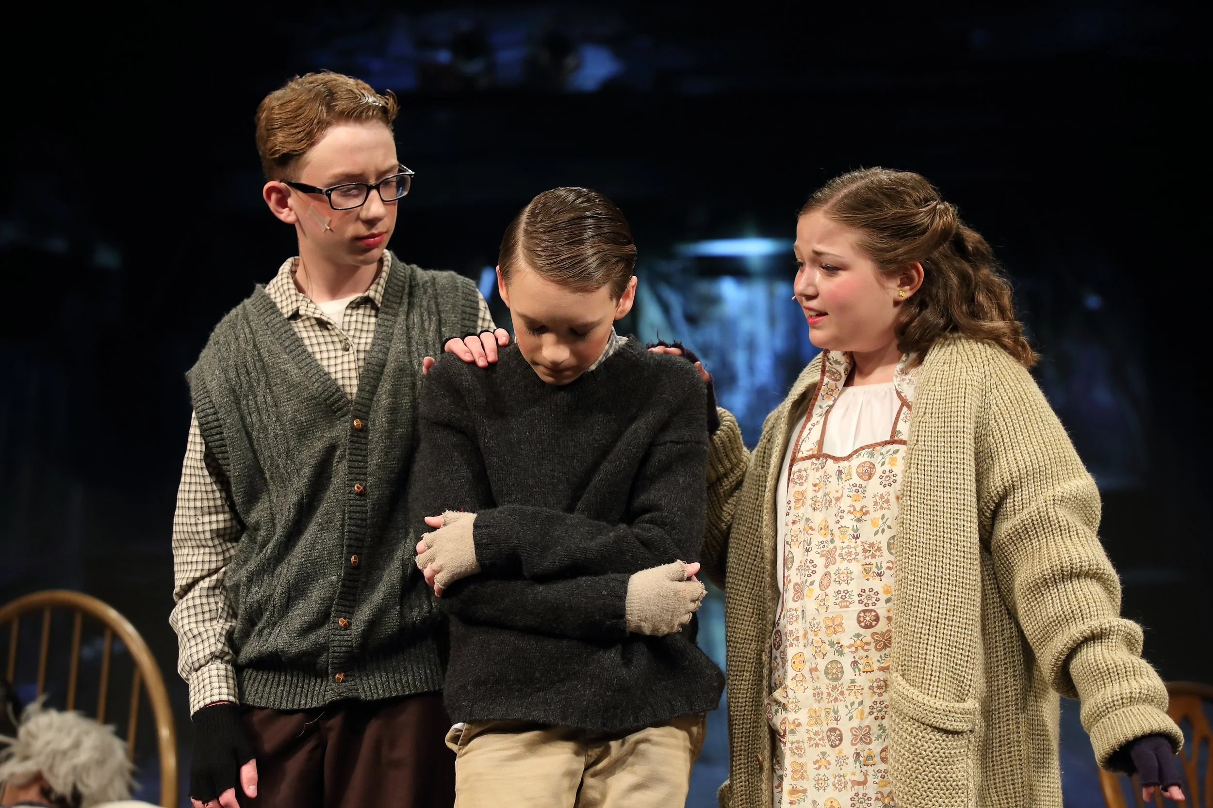 Three children standing close together, the boy in the middle has his head down and eyes closed, with a sad or tired expression, while the two children on either side look at him with concern. They are indoors with a dark background and a wooden chai