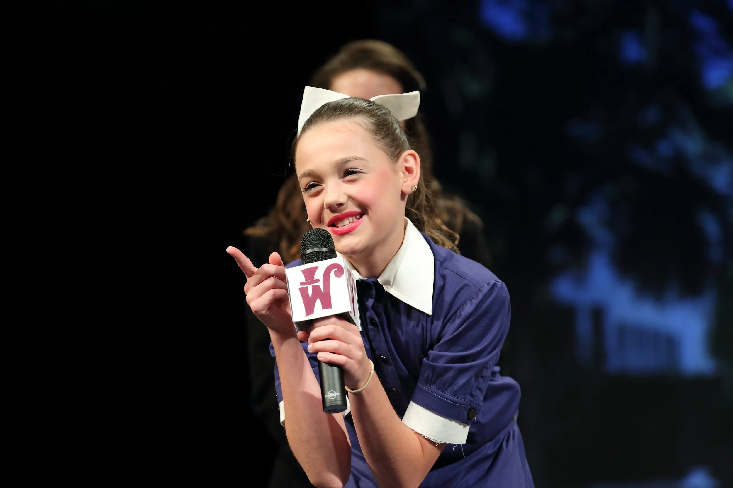 A young girl with a white bow in her hair, smiling and speaking into a microphone on stage, with another person partially visible behind her, blurred out.