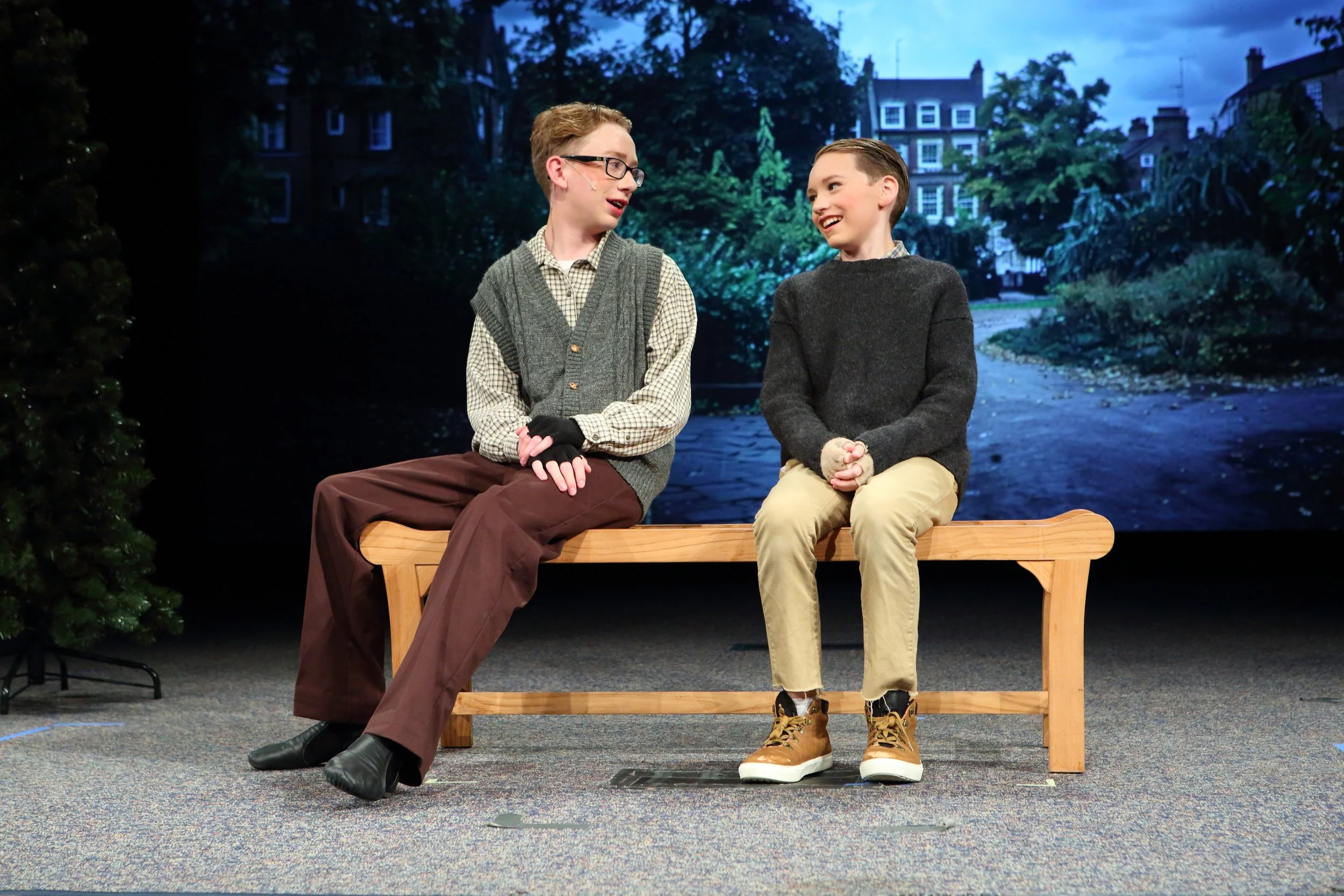 Two boys sitting on a wooden bench and smiling at each other. One boy wears glasses, a gray vest, checkered shirt, and brown pants. The other boy wears a dark gray sweater, beige pants, and tan boots. The background depicts a dark, outdoor scene with