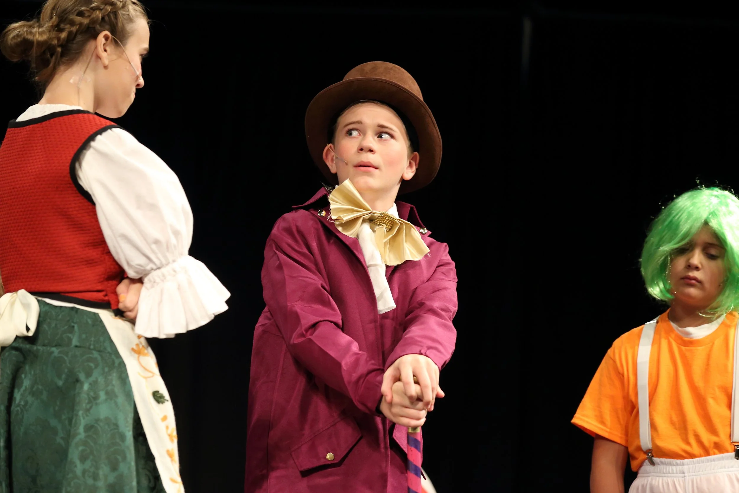 Three children in costume on stage, two girls and one boy in the center wearing a large brown hat and suspenders, with the girl on the left in period clothing and the girl on the right in a brightly colored costume with green hair.