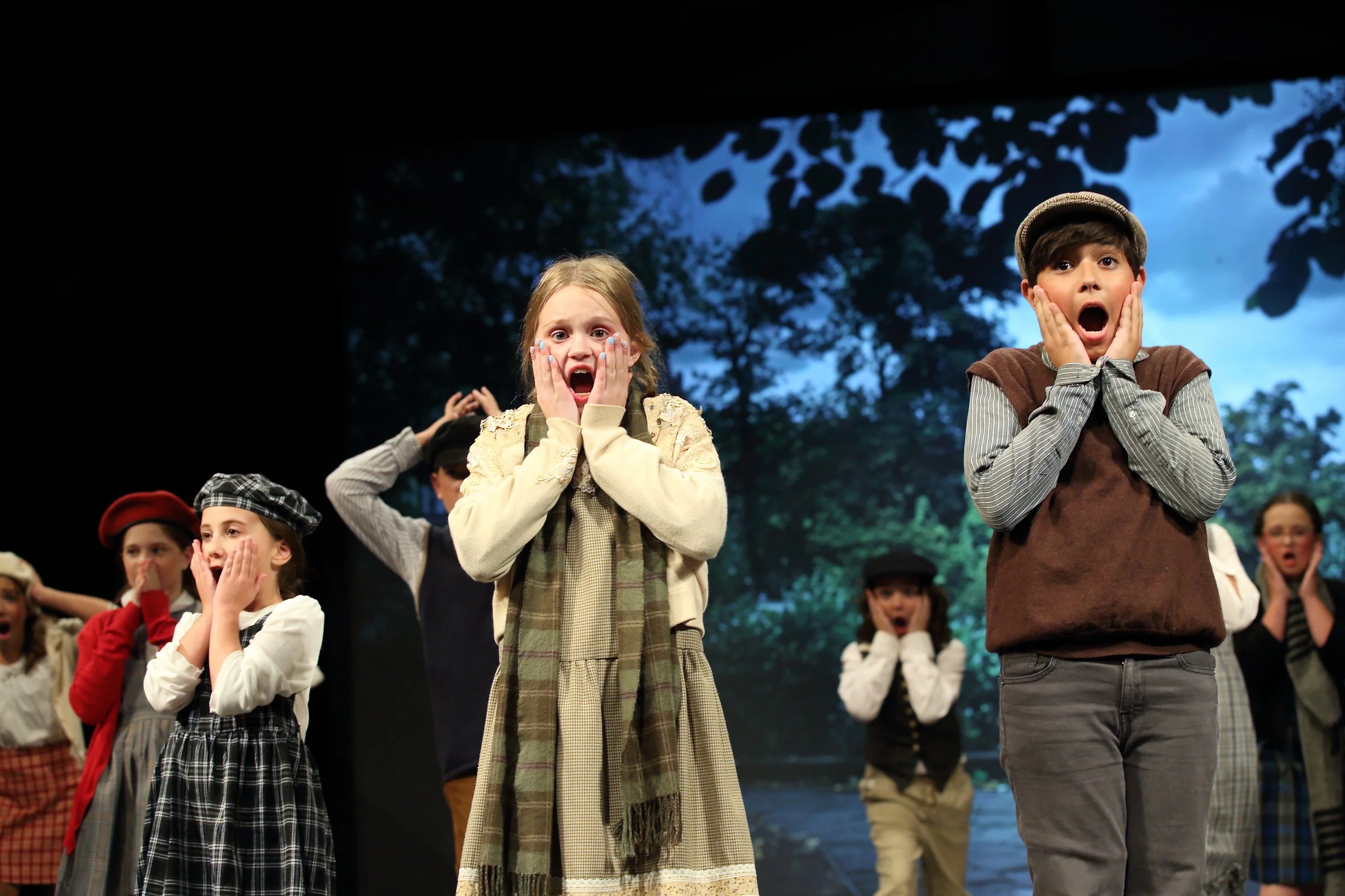Children dressed in early 20th-century clothing perform in a theatrical production, with expressions of shock or surprise, on a stage with a backdrop of trees and sky.
