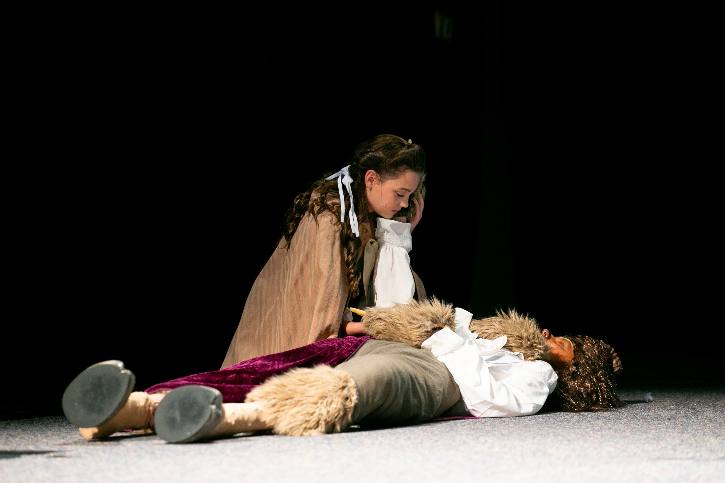 A girl with dark hair in braids and white ribbons, dressed in a historical costume, kneeling beside a person lying on the ground dressed in a costume with fur and gold details, in a theatrical performance with a black background.