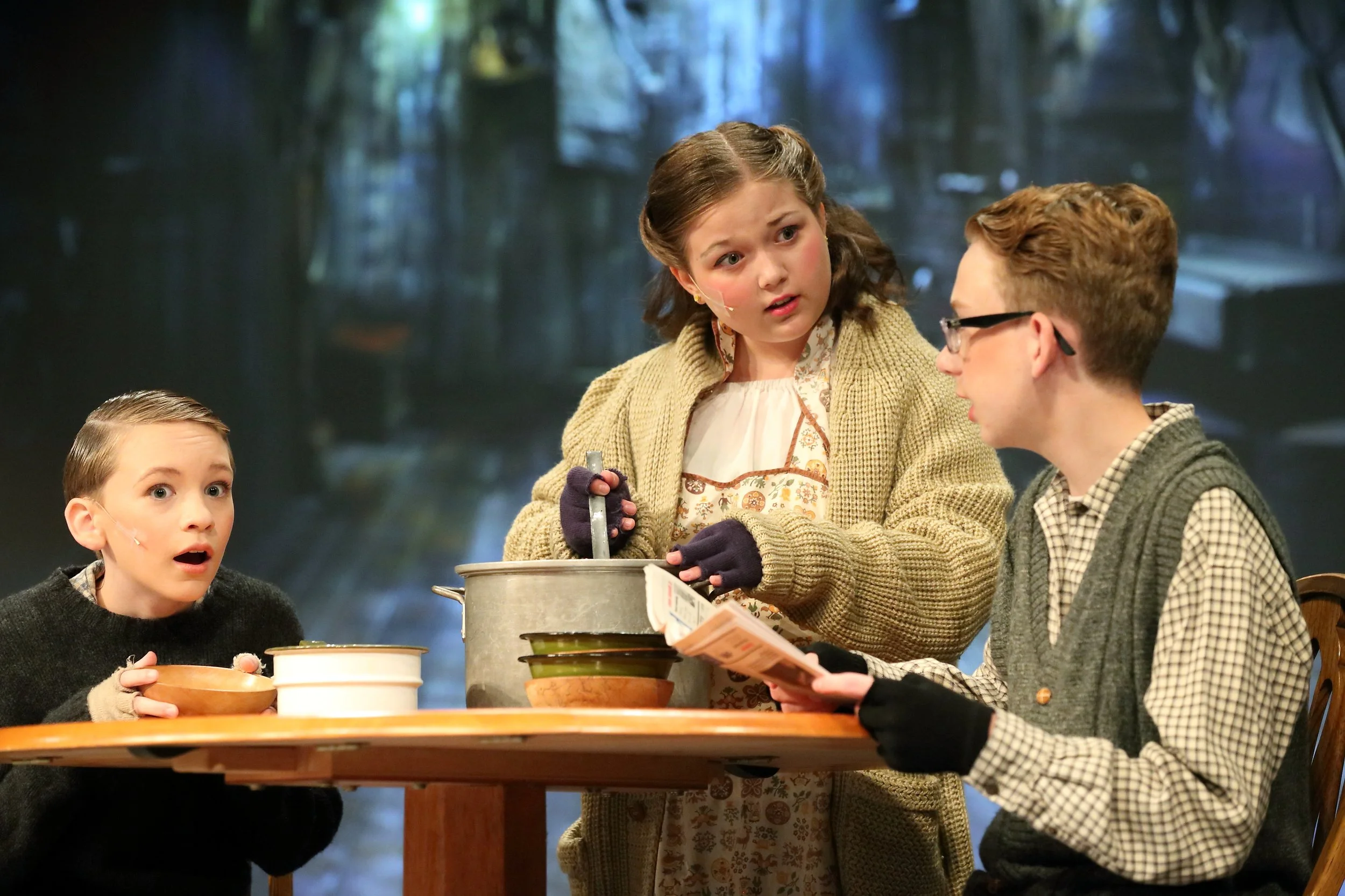 Four children are sitting at a wooden table with food containers, with a dark scenic background. One girl is holding a spoon over a large pot, and one boy is holding a paper, another is looking surprised, and the fourth girl is looking at the boy wit