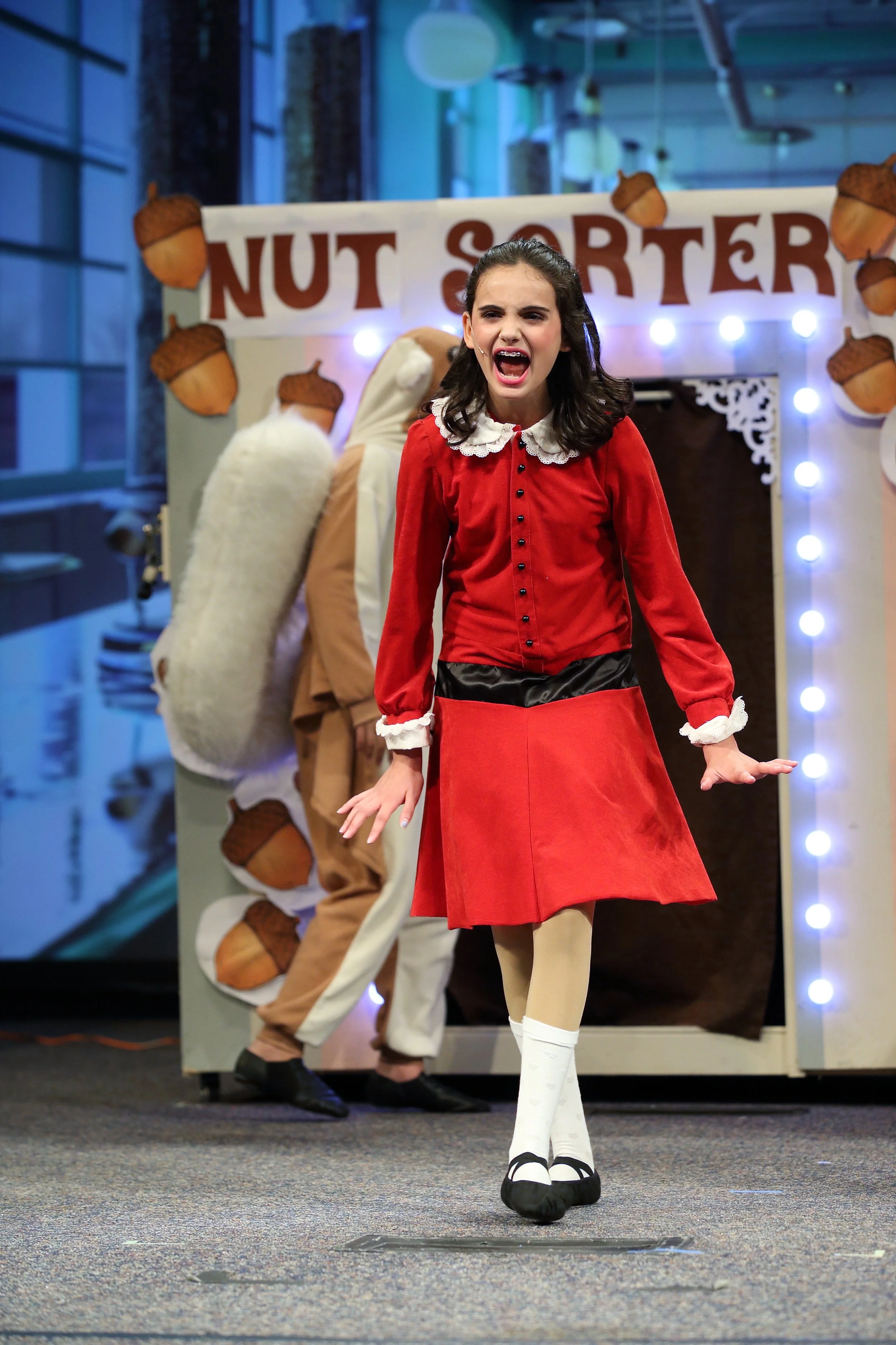 Young girl dressed as an elf or reindeer, performing on stage with a backing display that reads 'NUT SOR' and decorated with acorn cutouts. She appears to be singing or speaking with an expressive face, wearing a red dress, white tights, and black sh