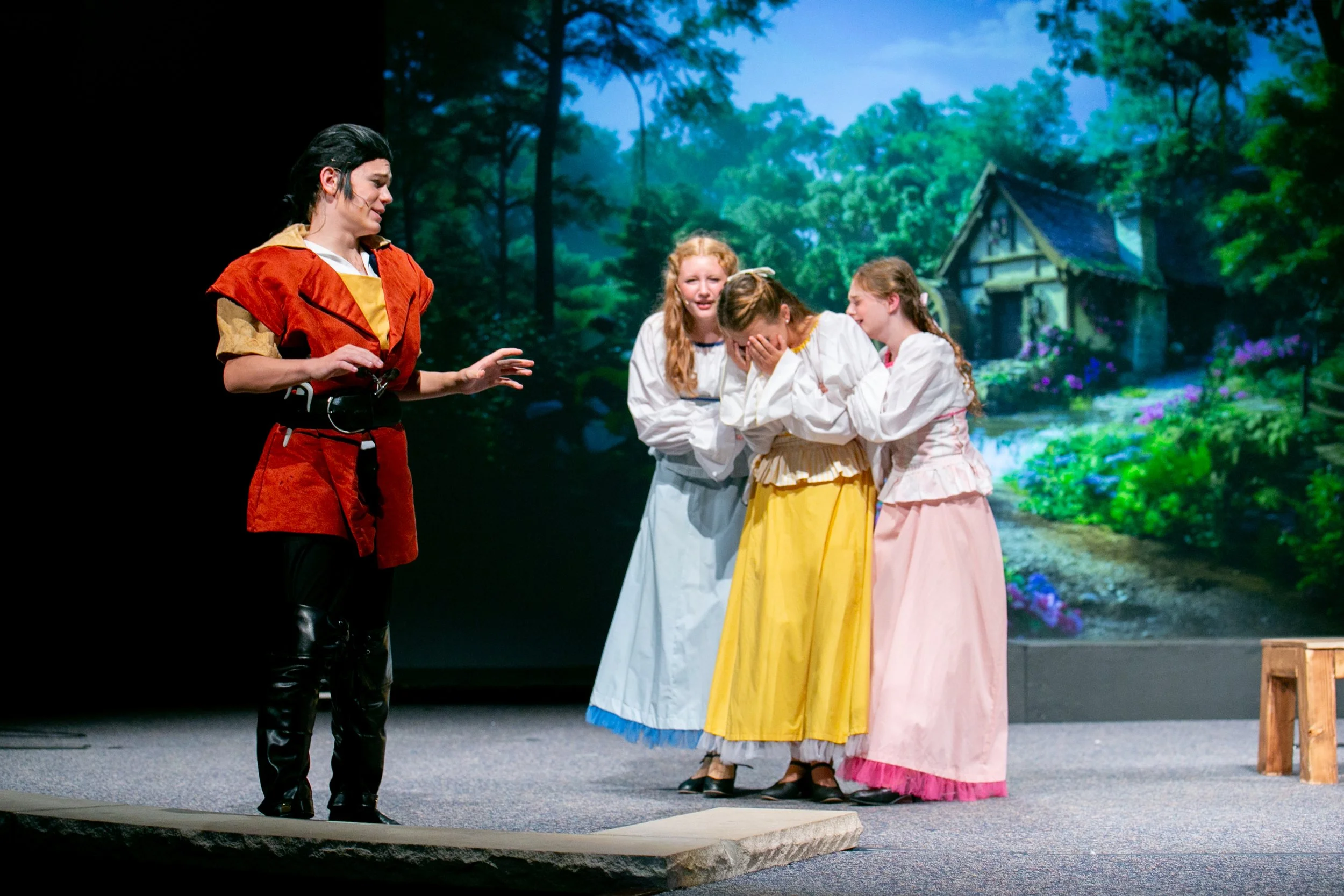 A theatrical scene with four girls dressed in historical clothing, one girl standing apart gesturing with her hands, and three girls reacting emotionally, set against a backdrop of a painted forest landscape with a house and stream.