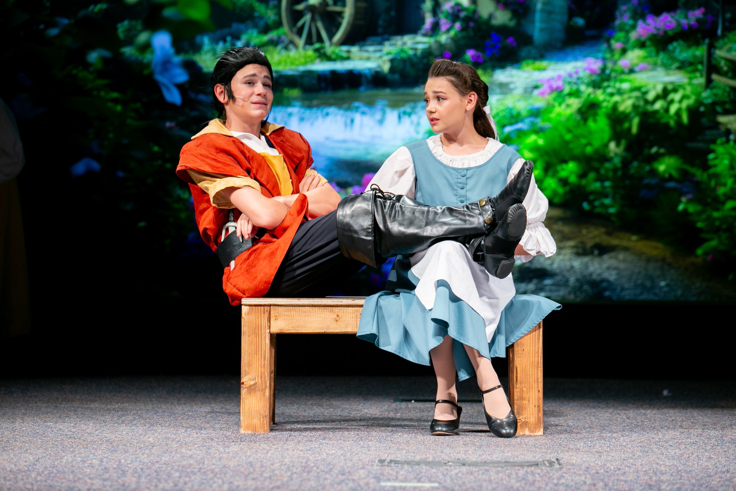 Stage scene with a young man and woman in costume, sitting on a wooden bench against a backdrop of a painted nature scene with a waterfall and purple flowers. The young man appears distressed, wearing a medieval or fantasy costume with a shoulder pat