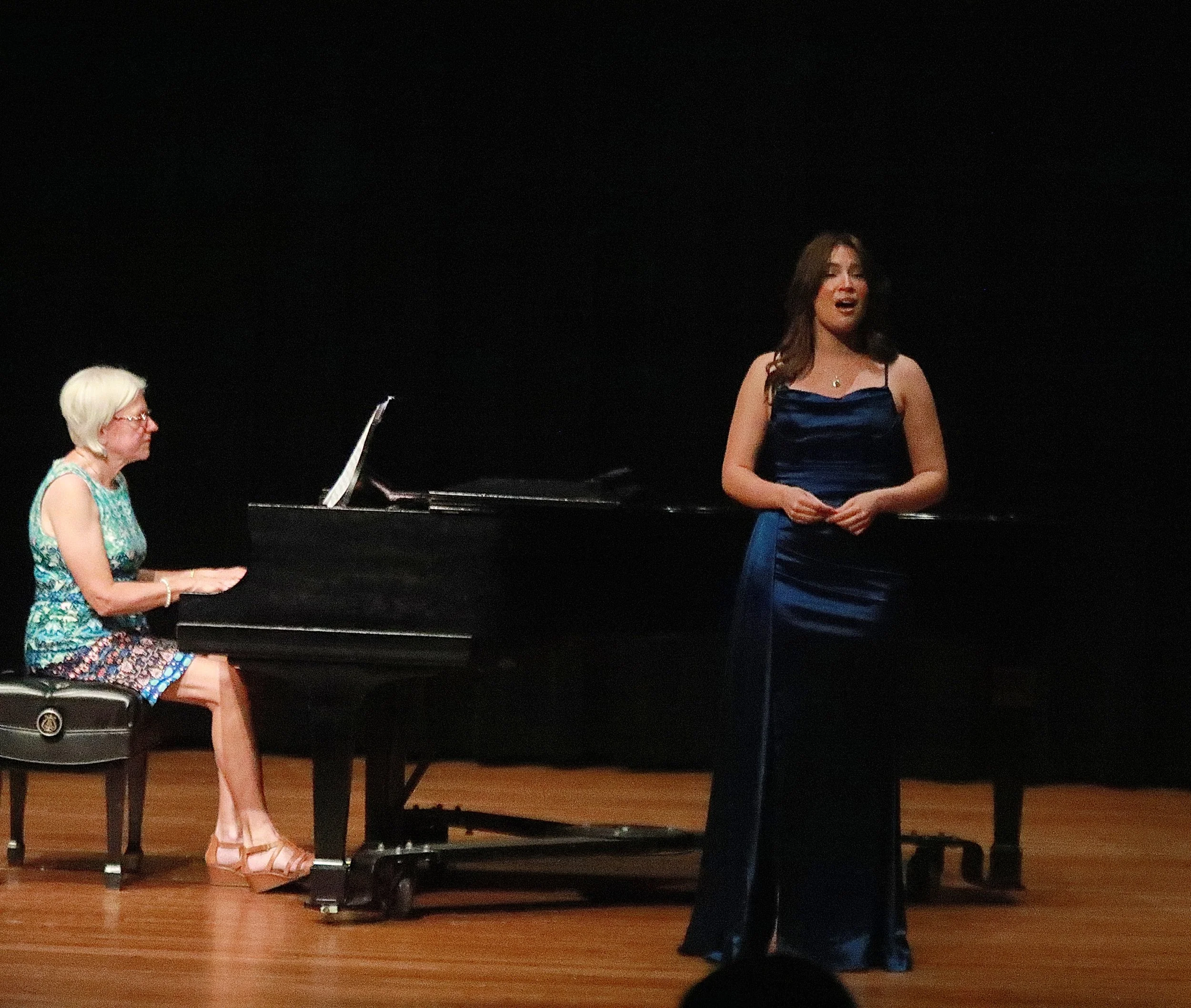 A woman in a blue dress standing near a woman playing the piano during a performance on stage.