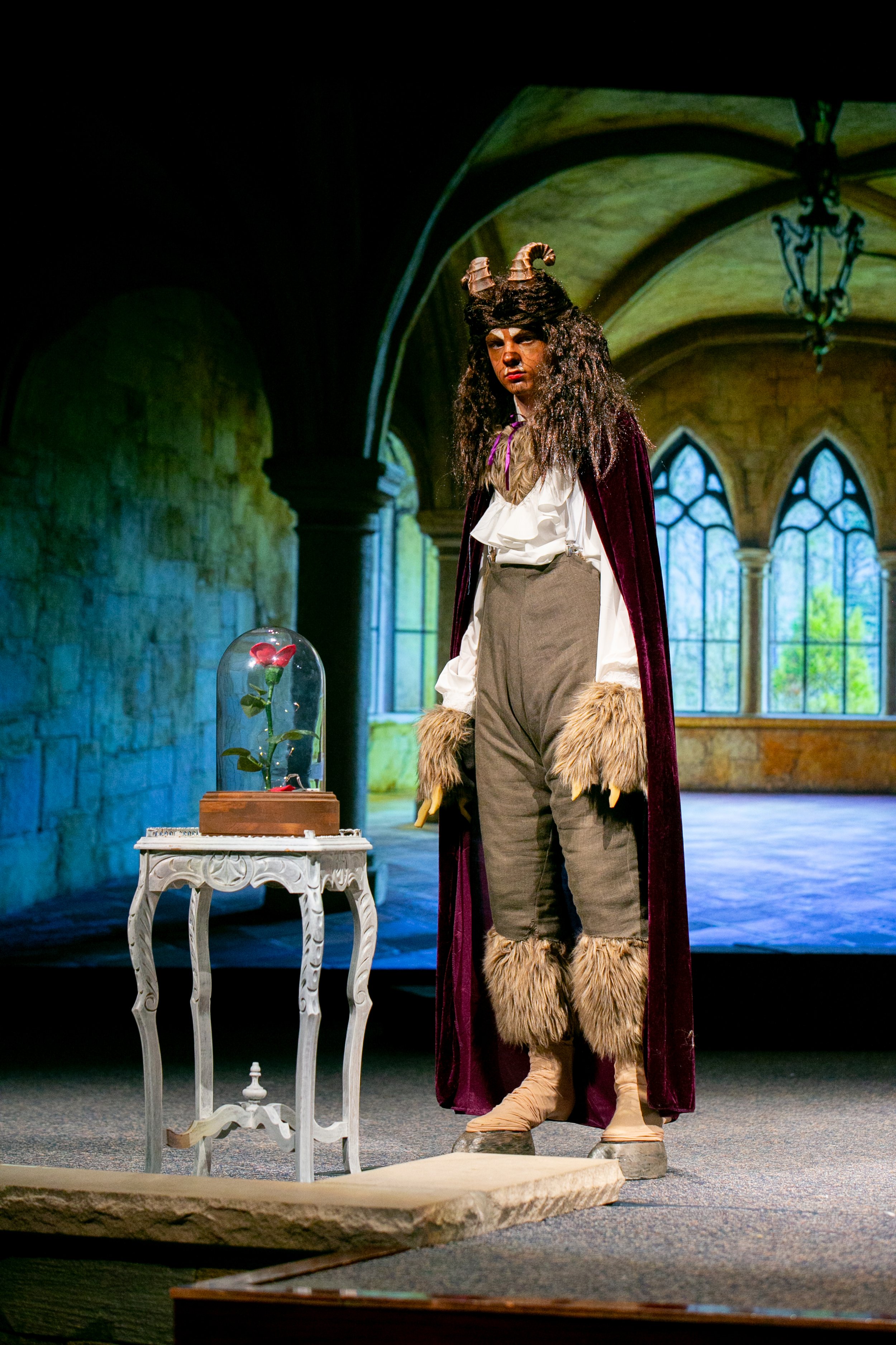 Person dressed in a costume resembling a mythical creature, standing in an indoor space with large arched windows, next to a white ornate table with a glass dome covering a red rose.