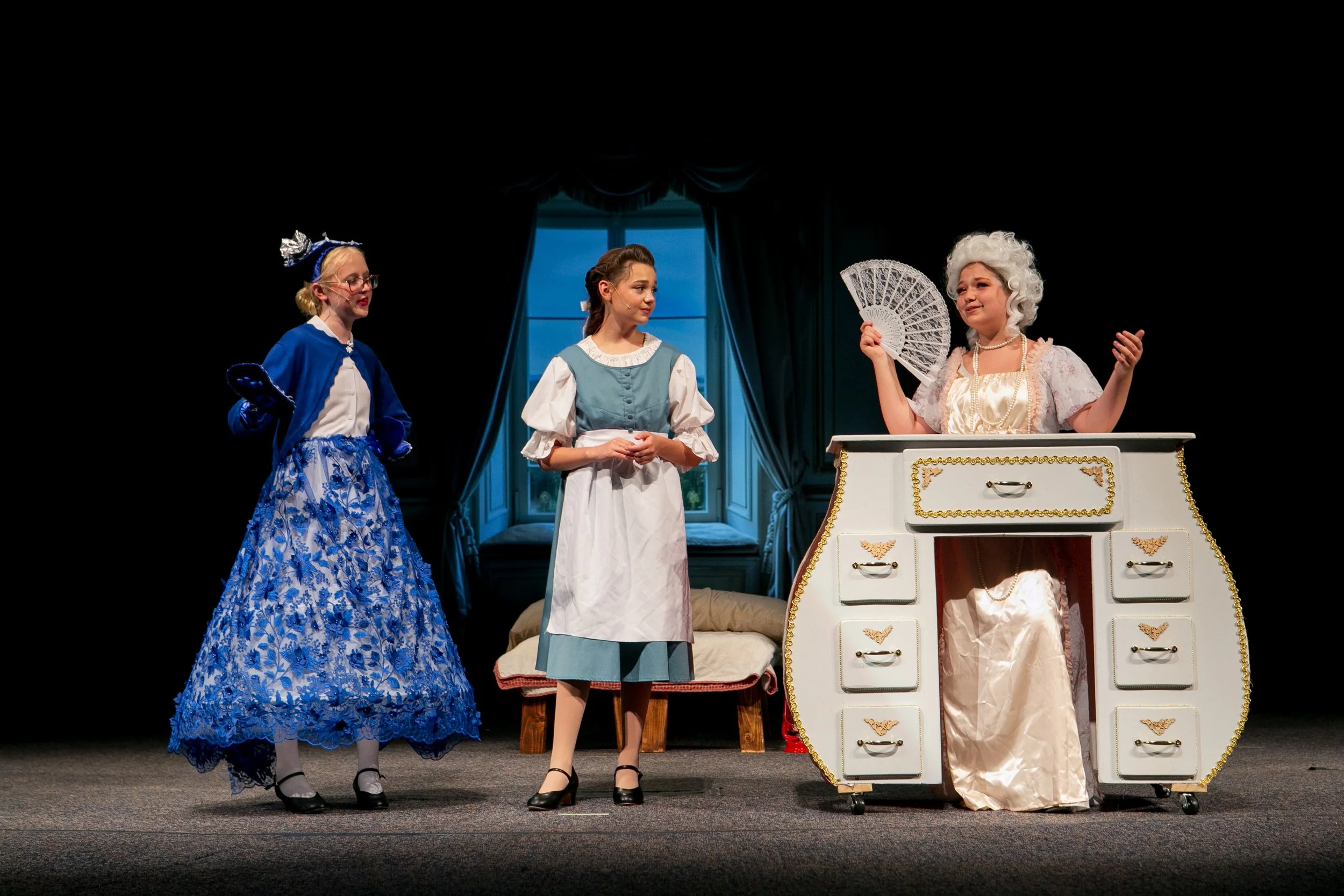 Three women dressed in period costumes perform on stage with a vintage dresser and a window in the background.