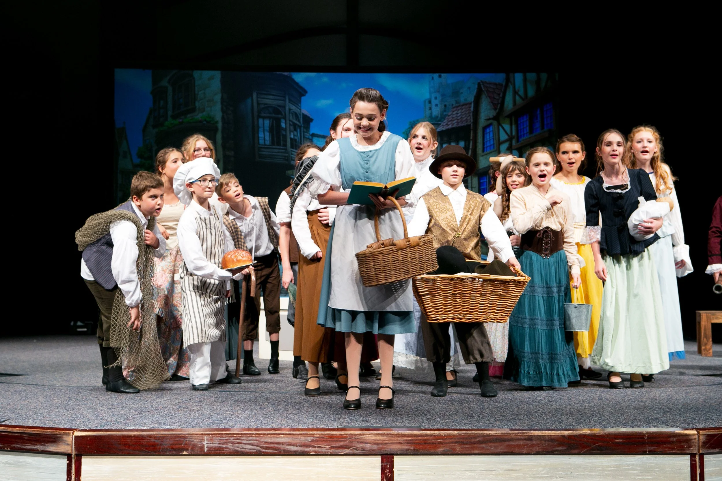 Children in costume performing on stage, portraying characters from a historical or fairy tale setting, with a backdrop of a village scene.