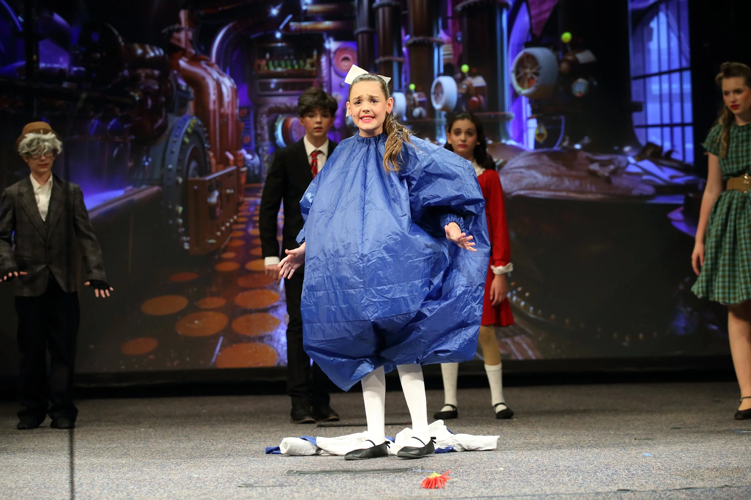 A girl dressed as a scarecrow performing on stage with a group of children in costume, a colorful set behind them.
