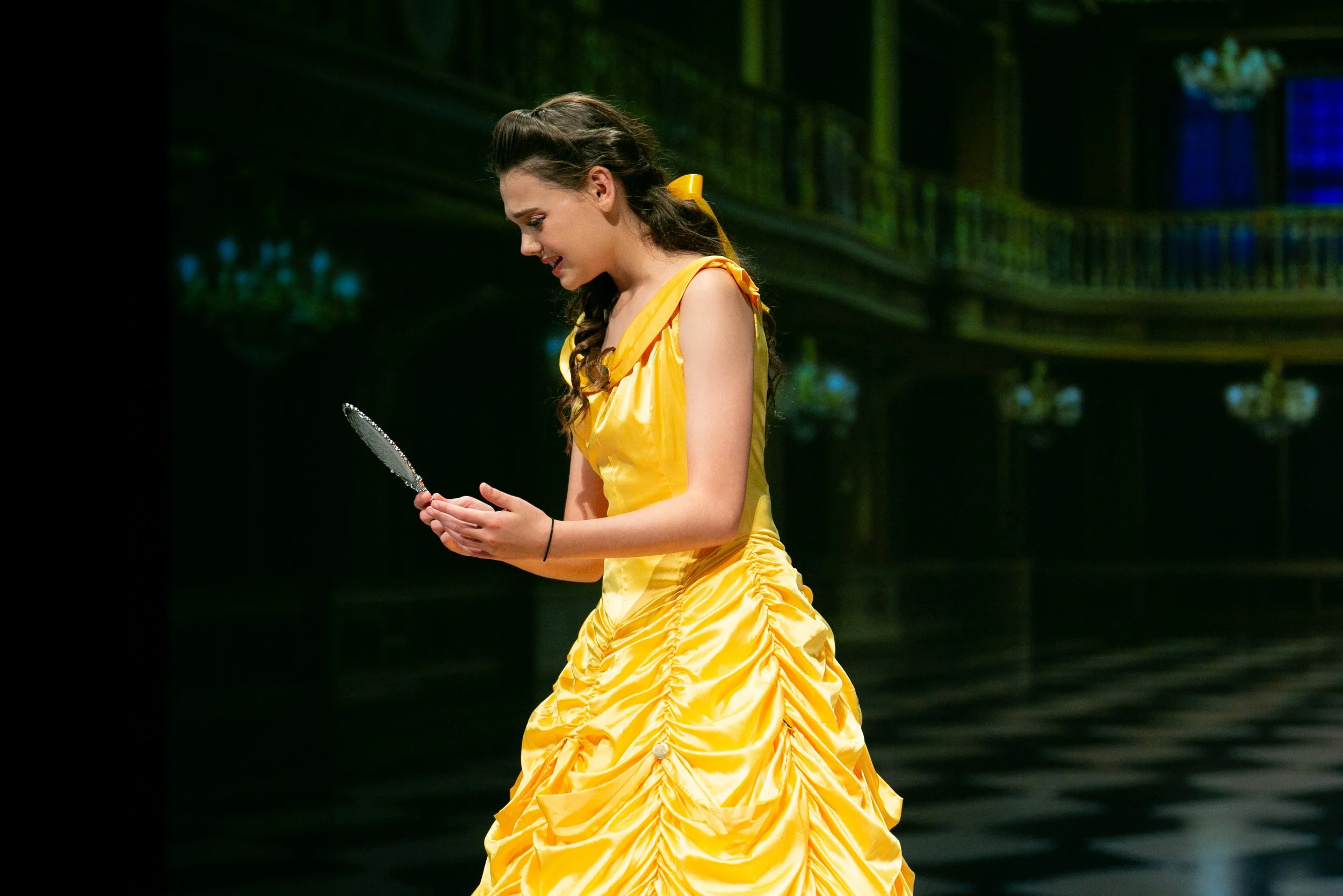 Young woman in a yellow dress holding a mirror and looking down, standing on a stage with dark background and green lighting.