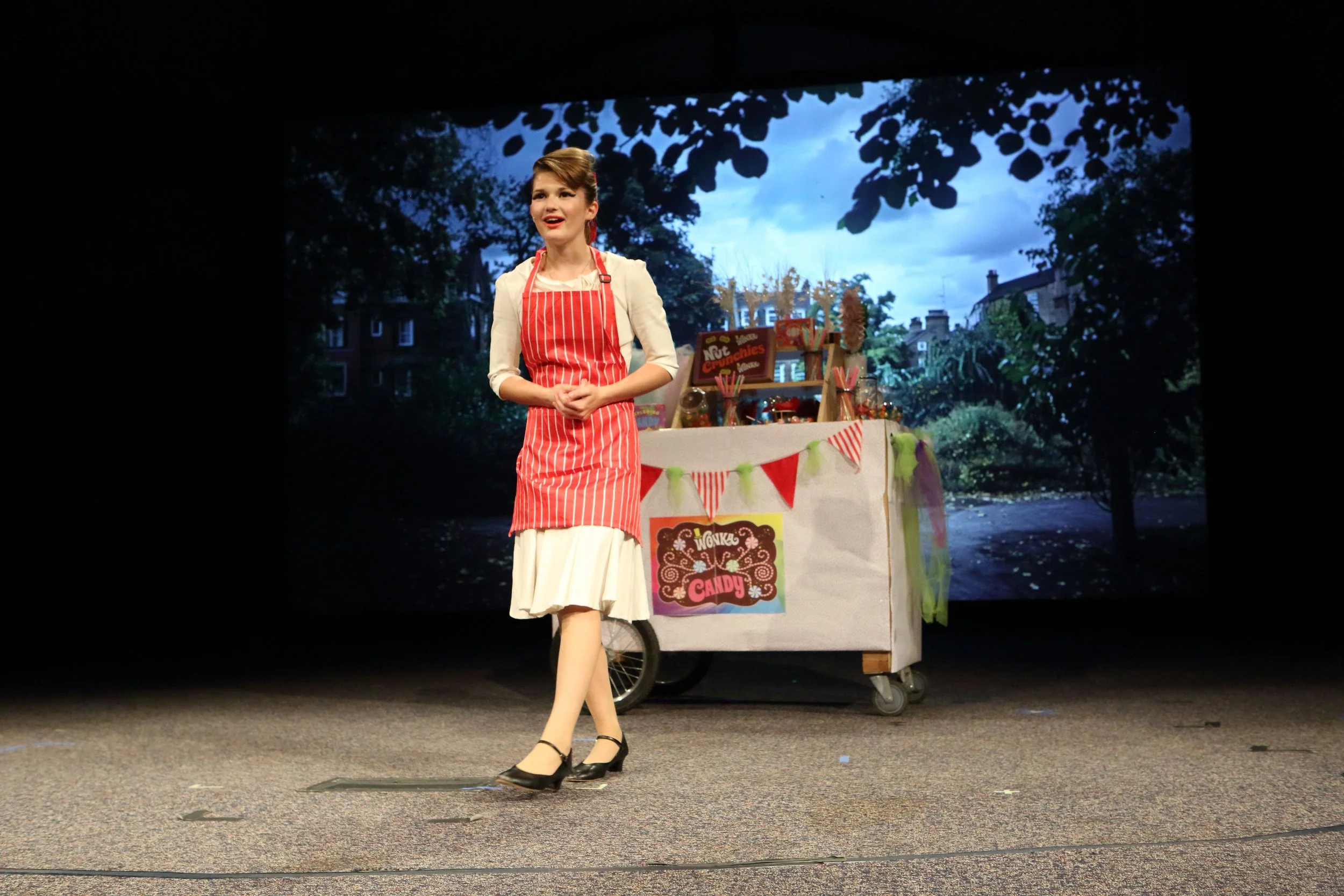 A woman walking on stage in front of a colorful candy stand backdrop, wearing a red and white striped apron and beige skirt, with a cloudy sky background visible behind her.