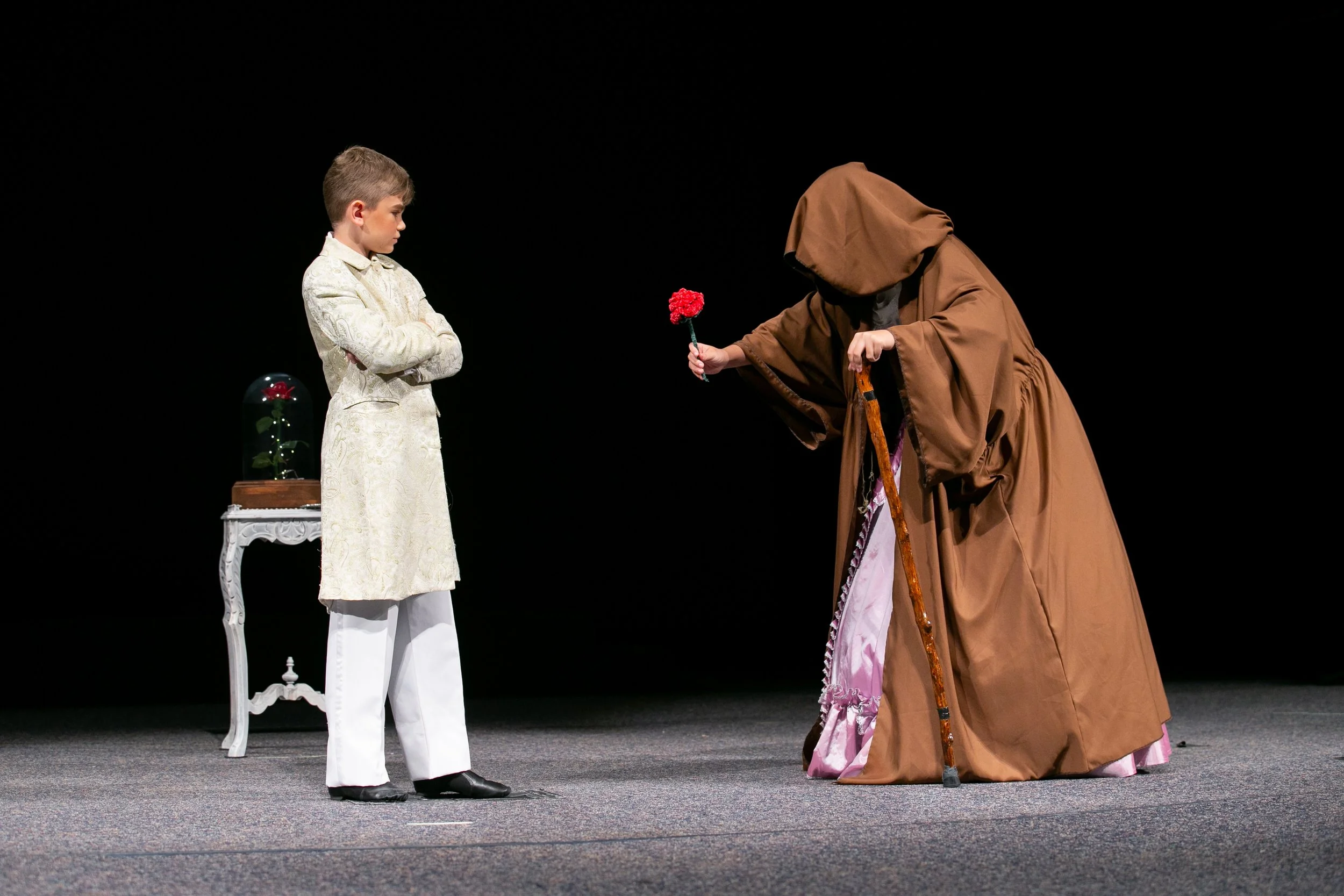 A young boy in a fancy white suit stands with arms crossed, facing an older person dressed as a wizard or wizard-like character who is handing him a red flower on a stage with a black background.