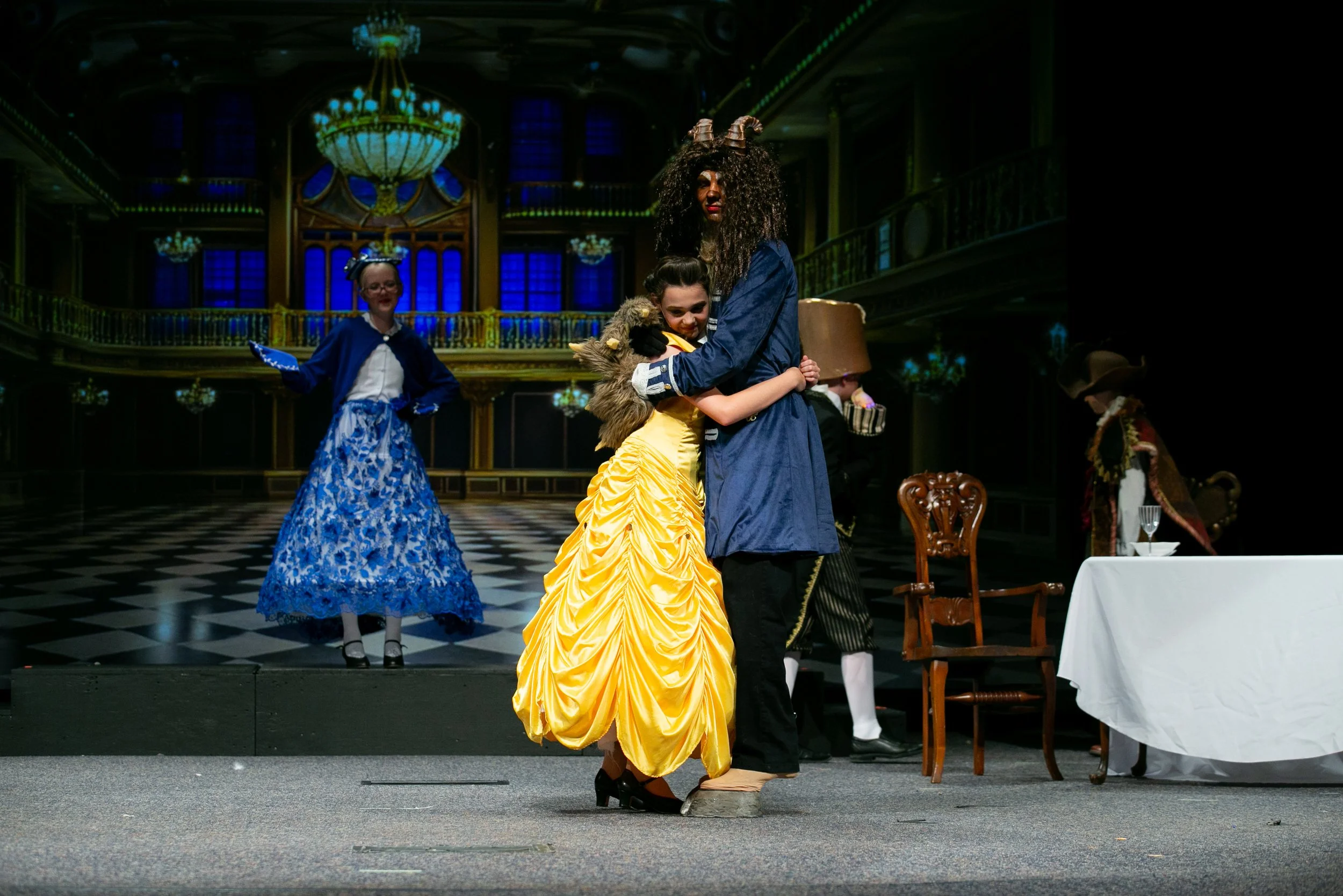 People dressed in elaborate costumes performing on stage in a grand theater with blue stained glass windows and ornate chandeliers.