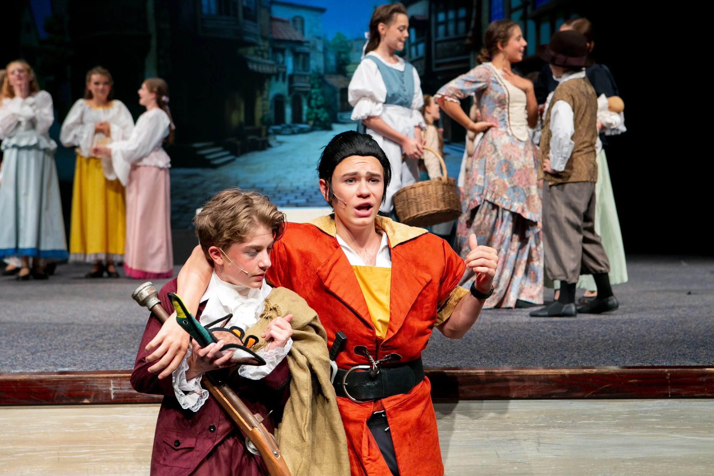 The image shows a scene from a theatrical play with children. Two children in the foreground are in costume, one with a sword and the other in an orange and yellow outfit, emotionally engaged. In the background, several children dressed in period cos