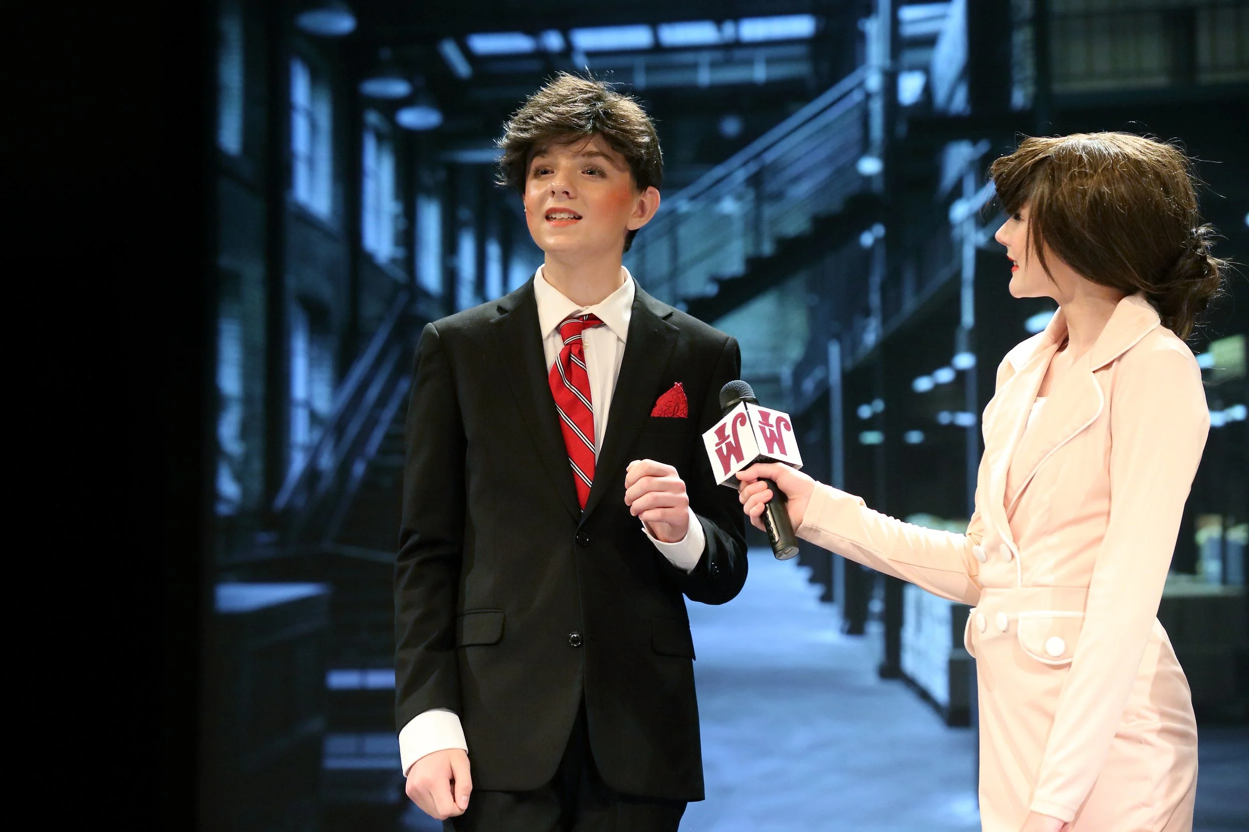 A young man in a suit and tie being interviewed by a woman holding a microphone that says 'EW'. They are standing outdoors with a modern building and stairs in the background.