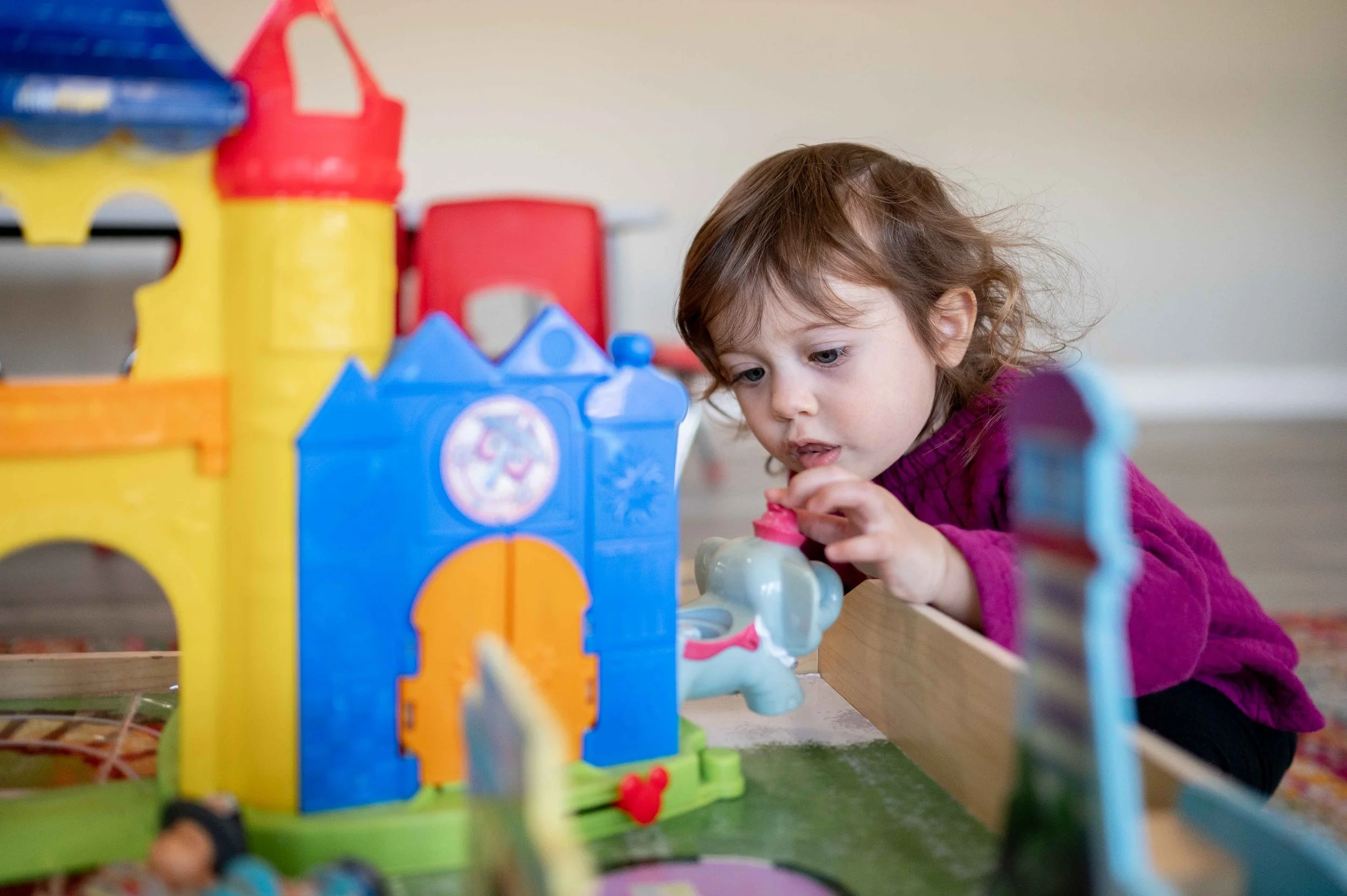 A young girl with brown hair in a purple sweater playing with colorful plastic building blocks on a table.