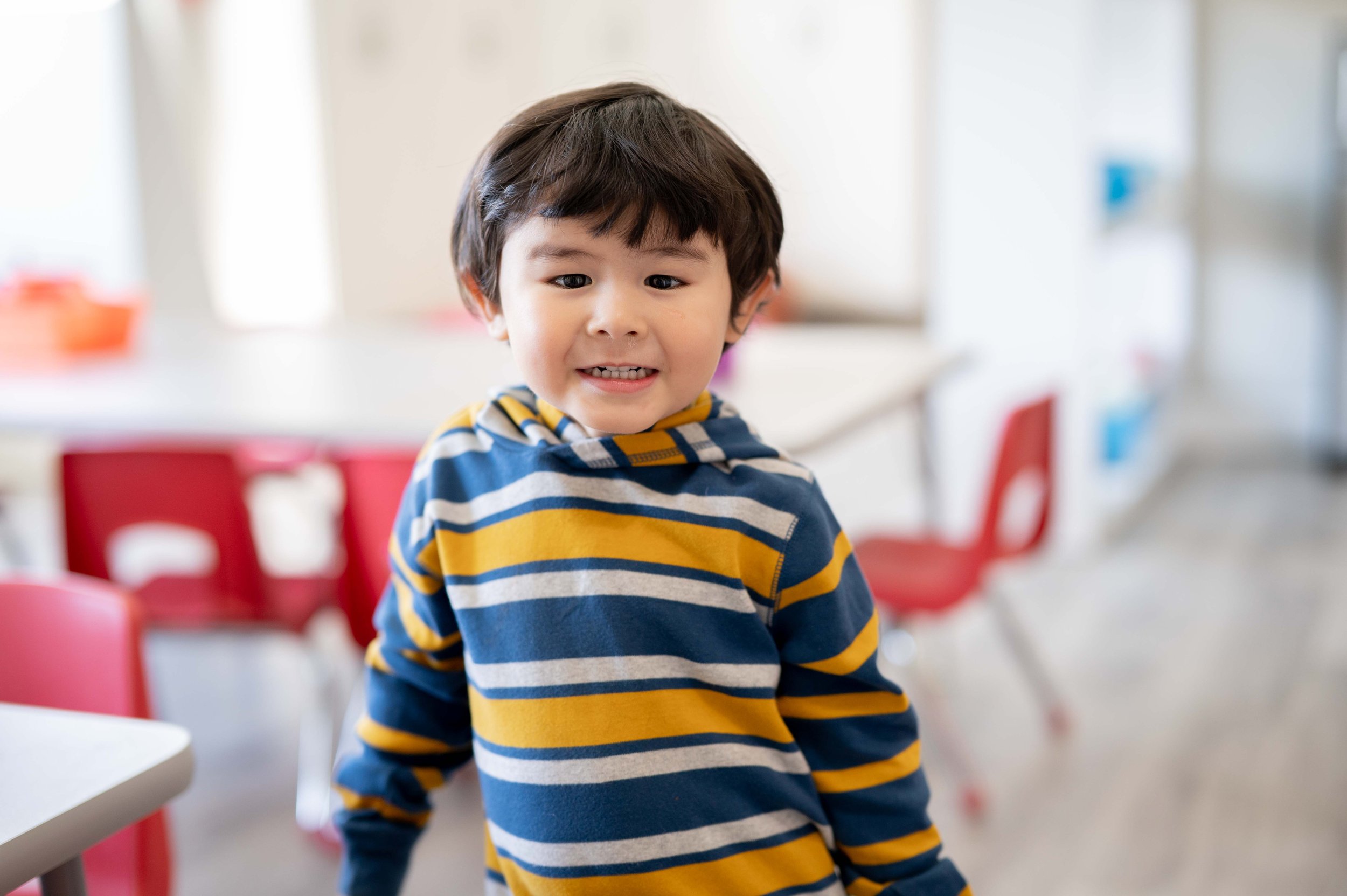 A young boy with dark hair and a striped yellow, blue, gray, and white shirt smiling at the camera in a bright room with red chairs and white tables.