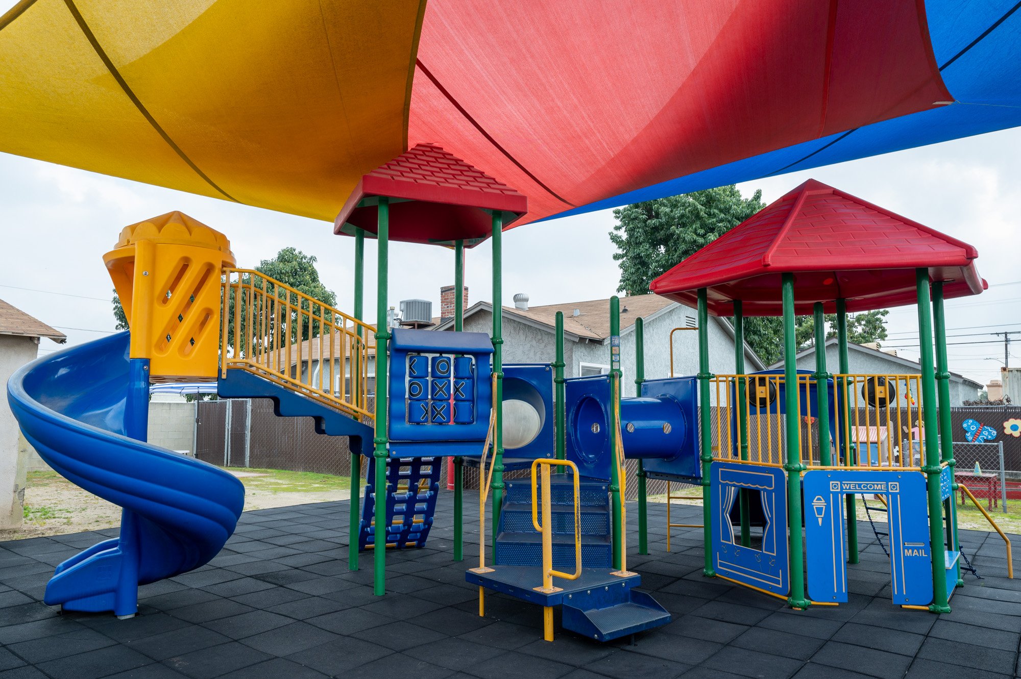Colorful outdoor playground with blue spiral slide, red and yellow shade canopies, and climbing structures, set on rubber flooring with residential houses and trees in the background.
