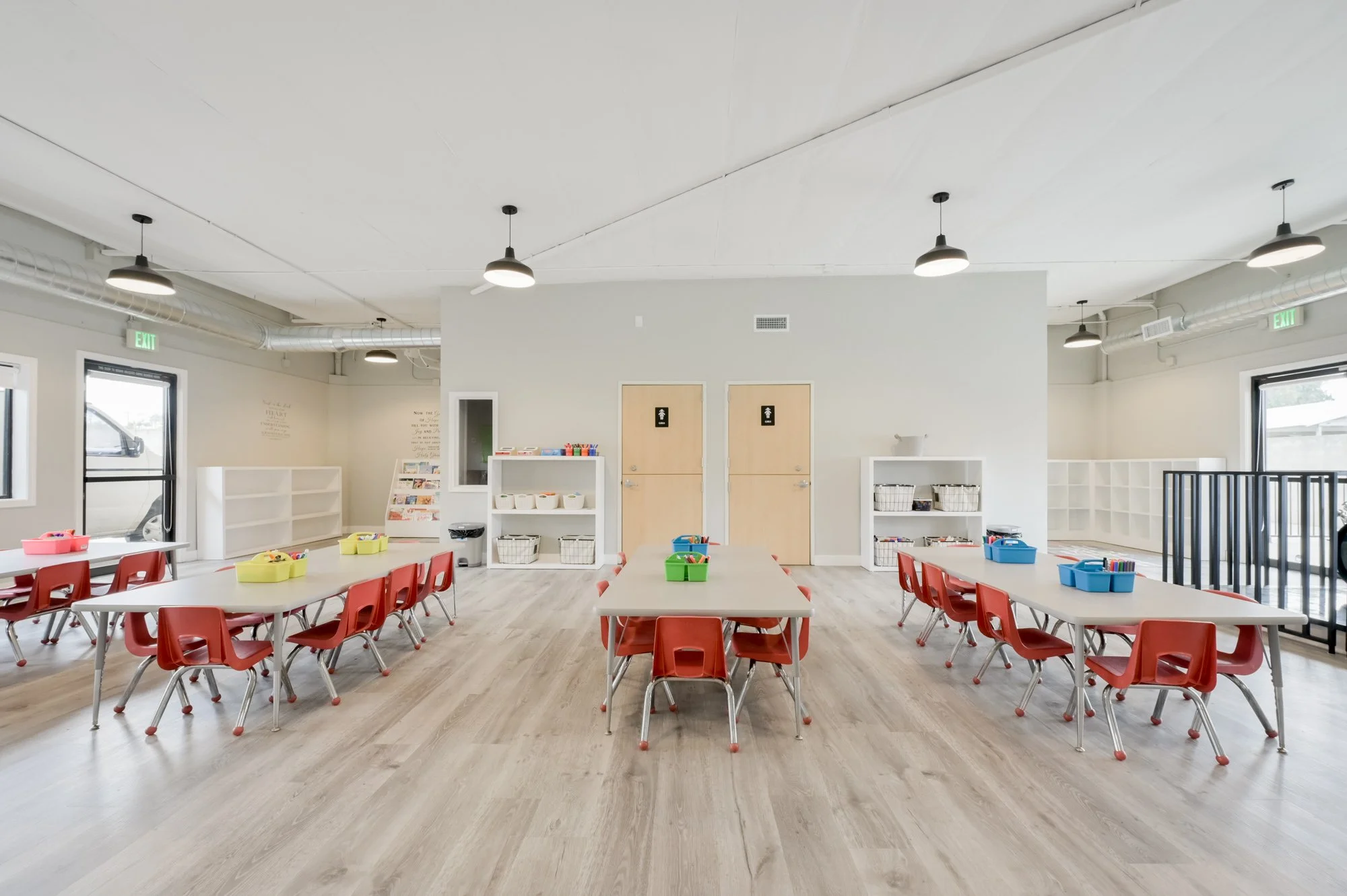 An empty classroom with light-colored flooring, white walls, and a white ceiling with multiple black pendant lights. There are red chairs and white tables arranged for children, with colorful bins for supplies. The room has large windows, exit signs,