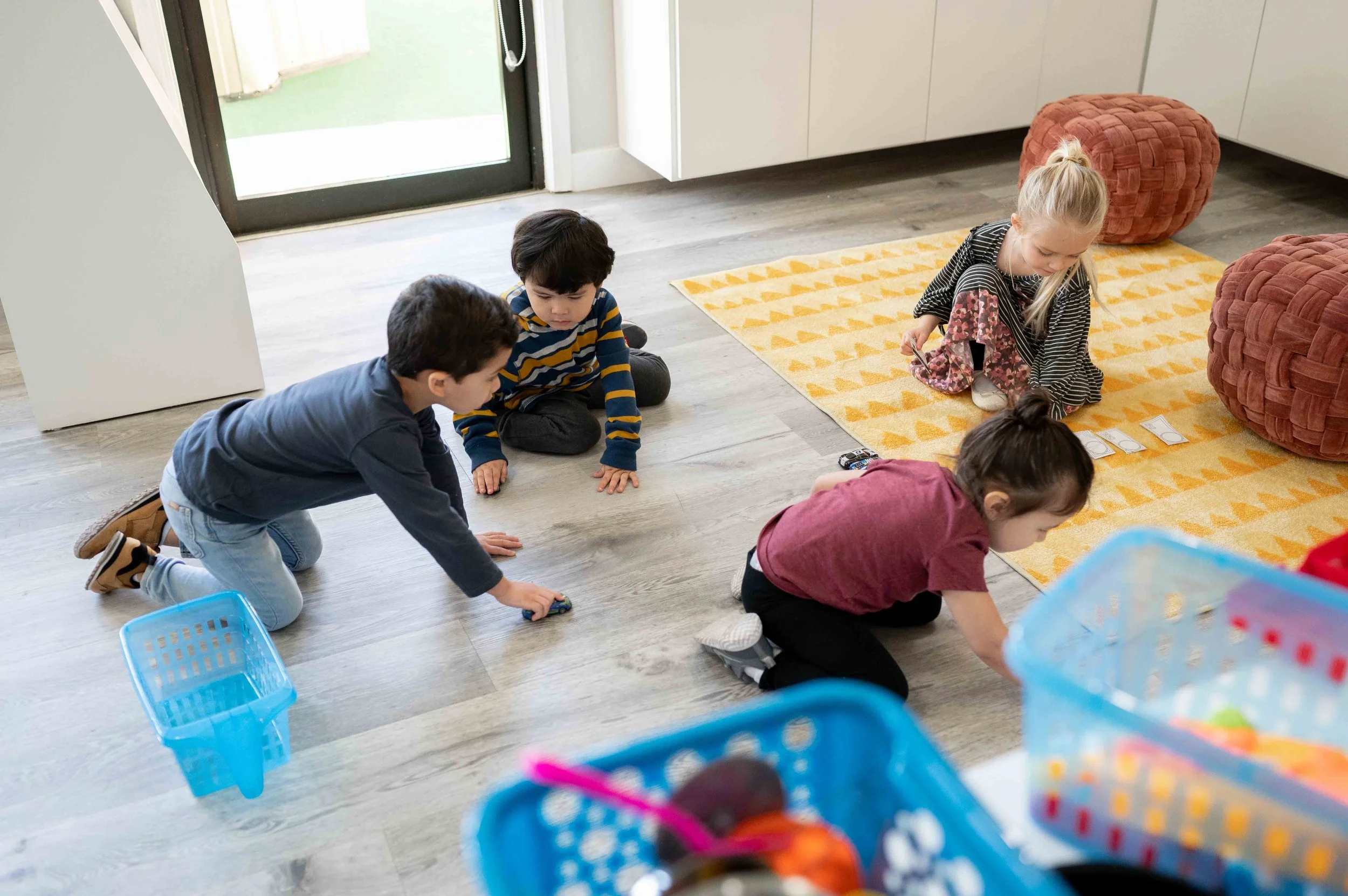 Children playing on the floor inside a house near a window with a door, some are kneeling and others are sitting, engaging with toys on a yellow rug with orange triangles.