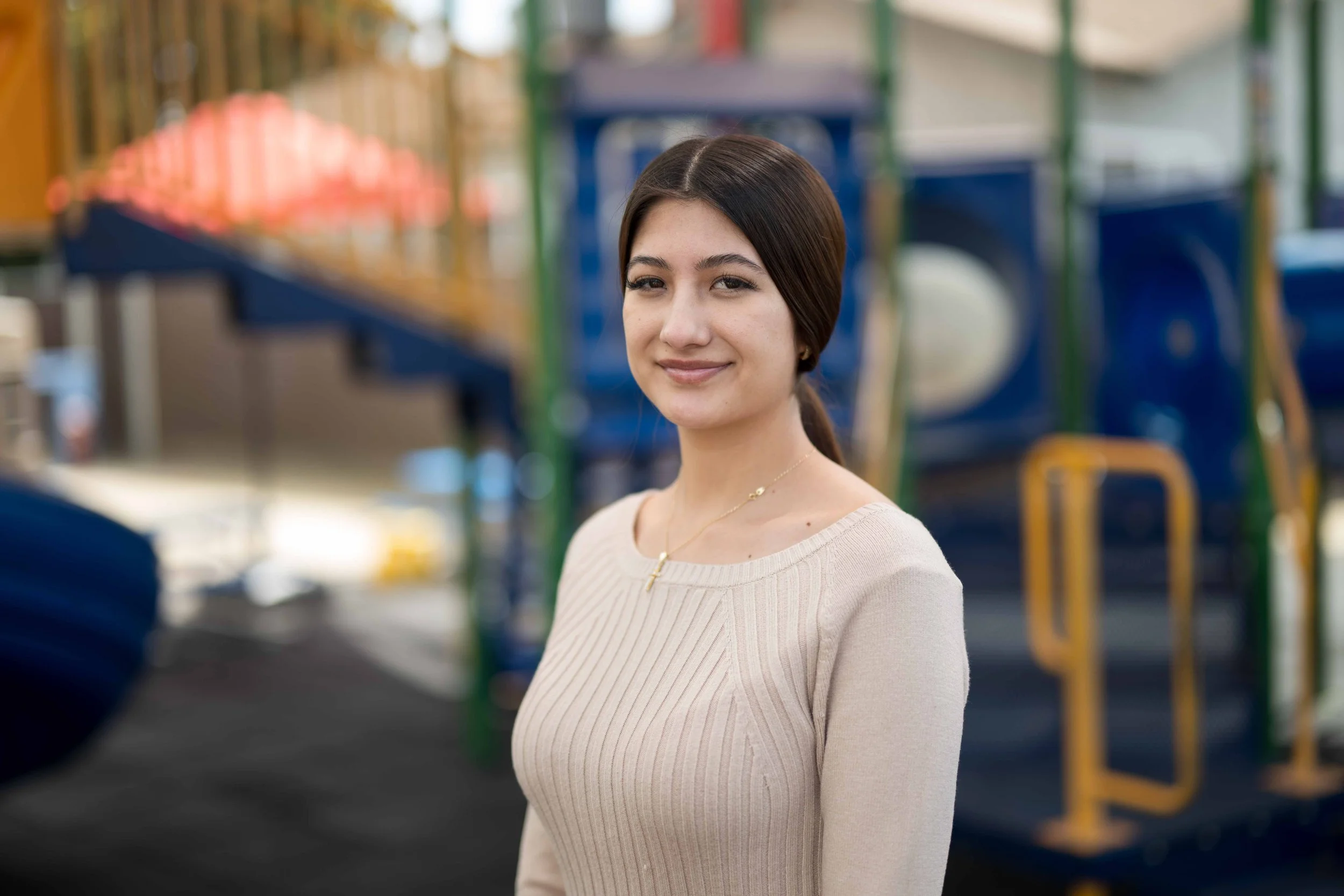 A young woman with dark brown hair smiling at the camera, standing in an outdoor playground area with colorful equipment blurred in the background.